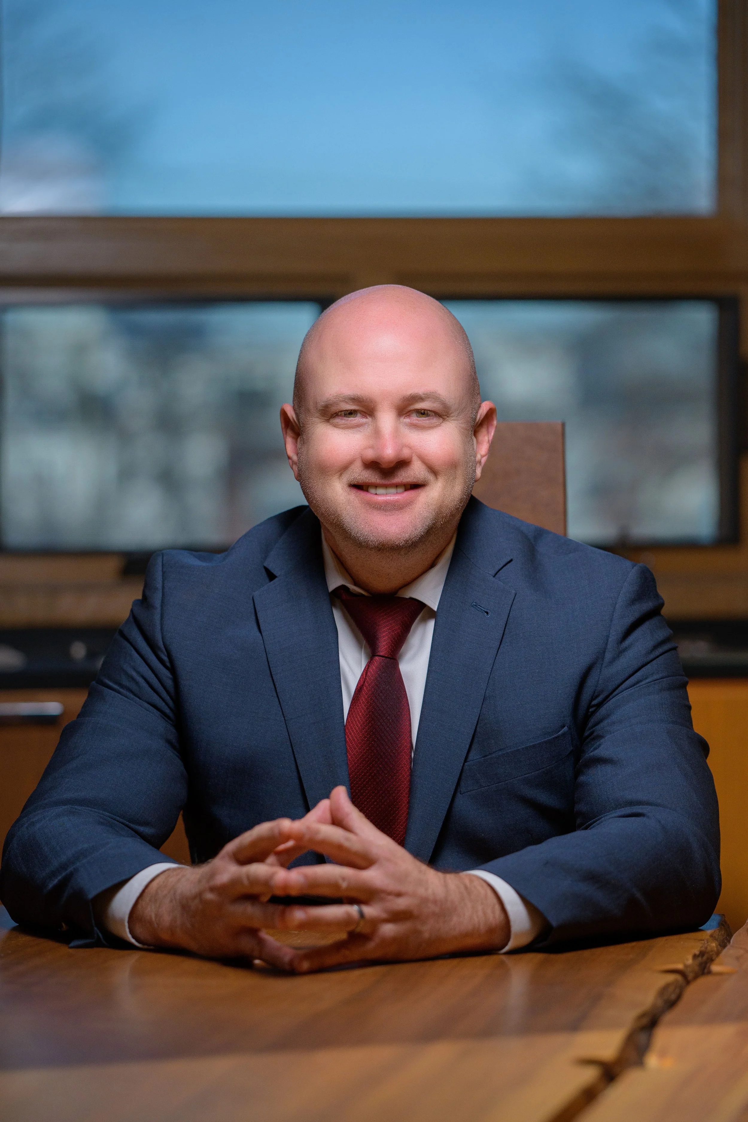 A man with a bald head, wearing a dark blue suit, white shirt, and maroon tie, sitting at a wooden table with his hands clasped in front of him. Behind him are large windows with a blurred outdoor scene.