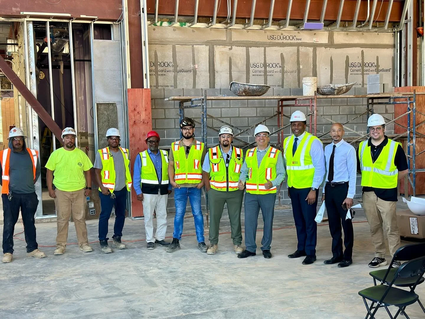 Group of construction workers and professionals in safety vests and helmets standing on a construction site with a partially built wall and scaffolding behind them.