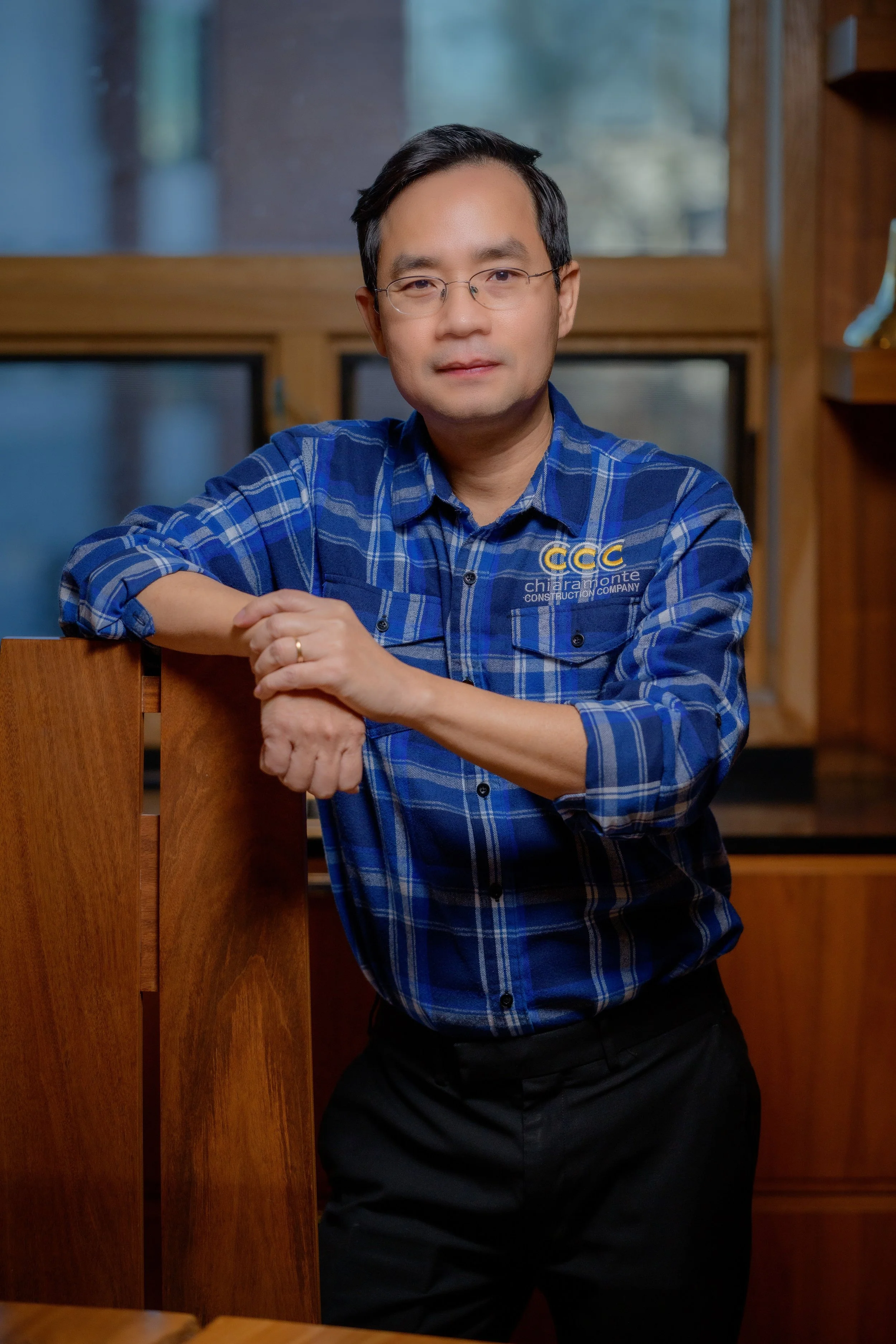 A man with glasses and short dark hair, wearing a blue checkered shirt with a logo, leaning against a wooden surface indoors.