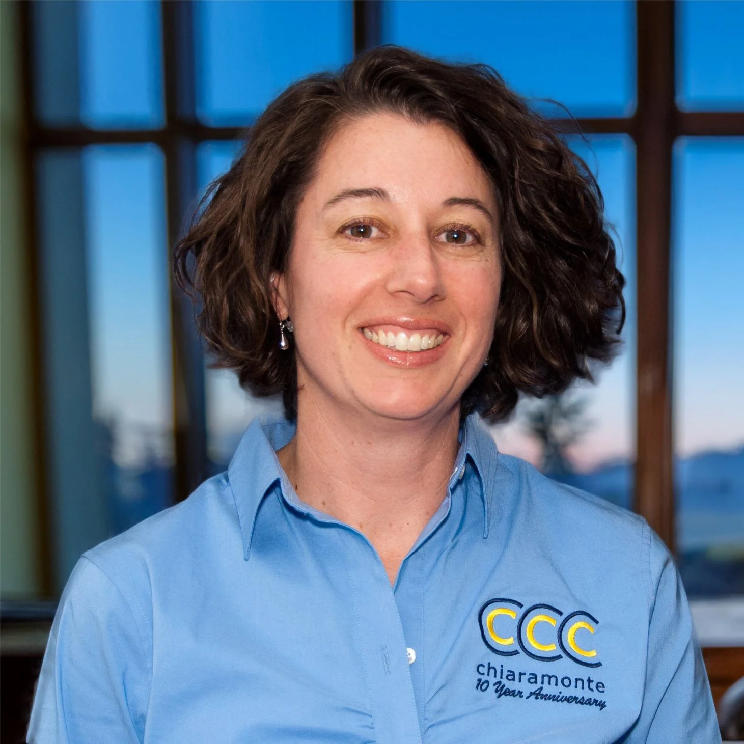 A smiling woman with short, curly brown hair wearing a blue shirt with a logo that reads 'CCC Chiaramonte 10 Year Anniversary,' standing in front of a window with a scenic view of the sunset.