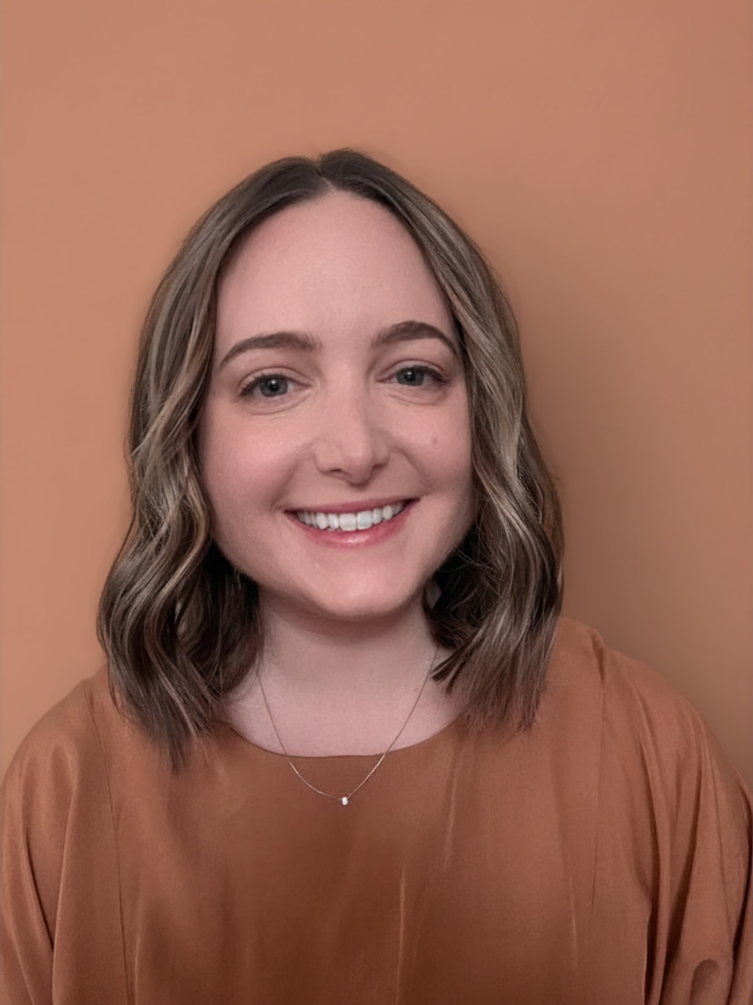 A young woman with shoulder-length wavy hair and blue eyes, smiling, wearing a brown top and a delicate necklace, against a plain beige background.