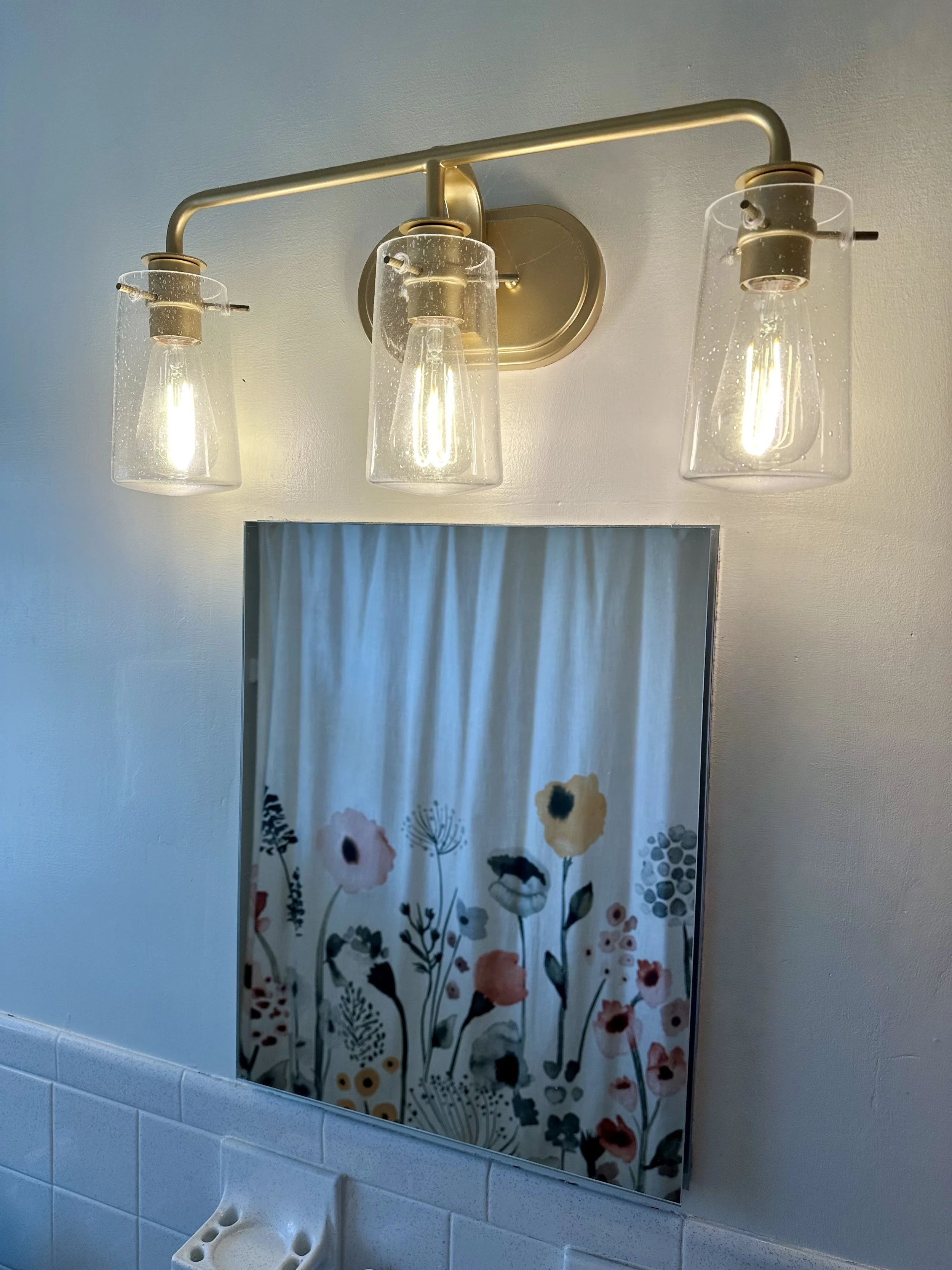 A bathroom wall mirror with a floral curtain reflected in it, above a white sink.