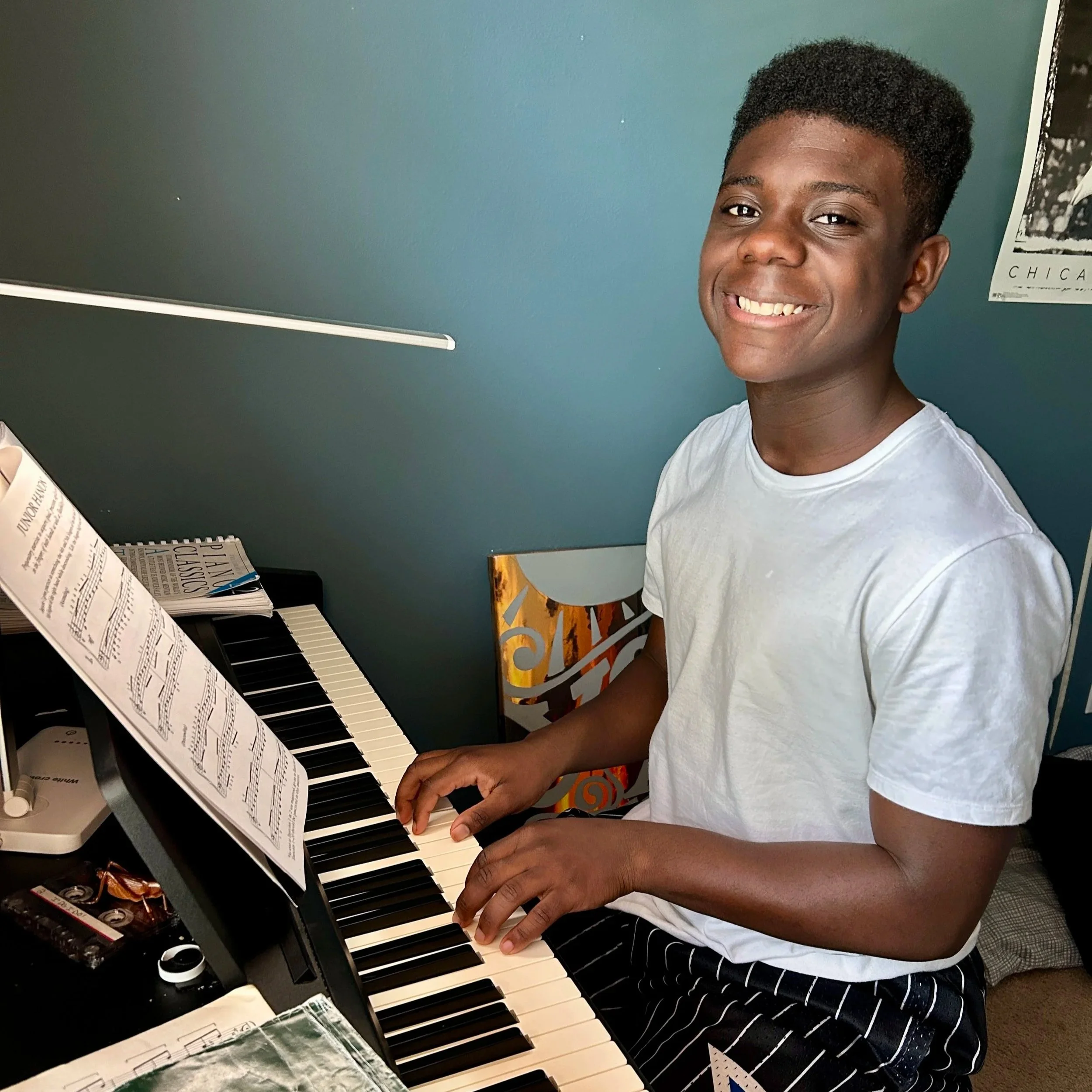 A young man sitting at a piano, smiling and playing music, with sheet music open in front of him.