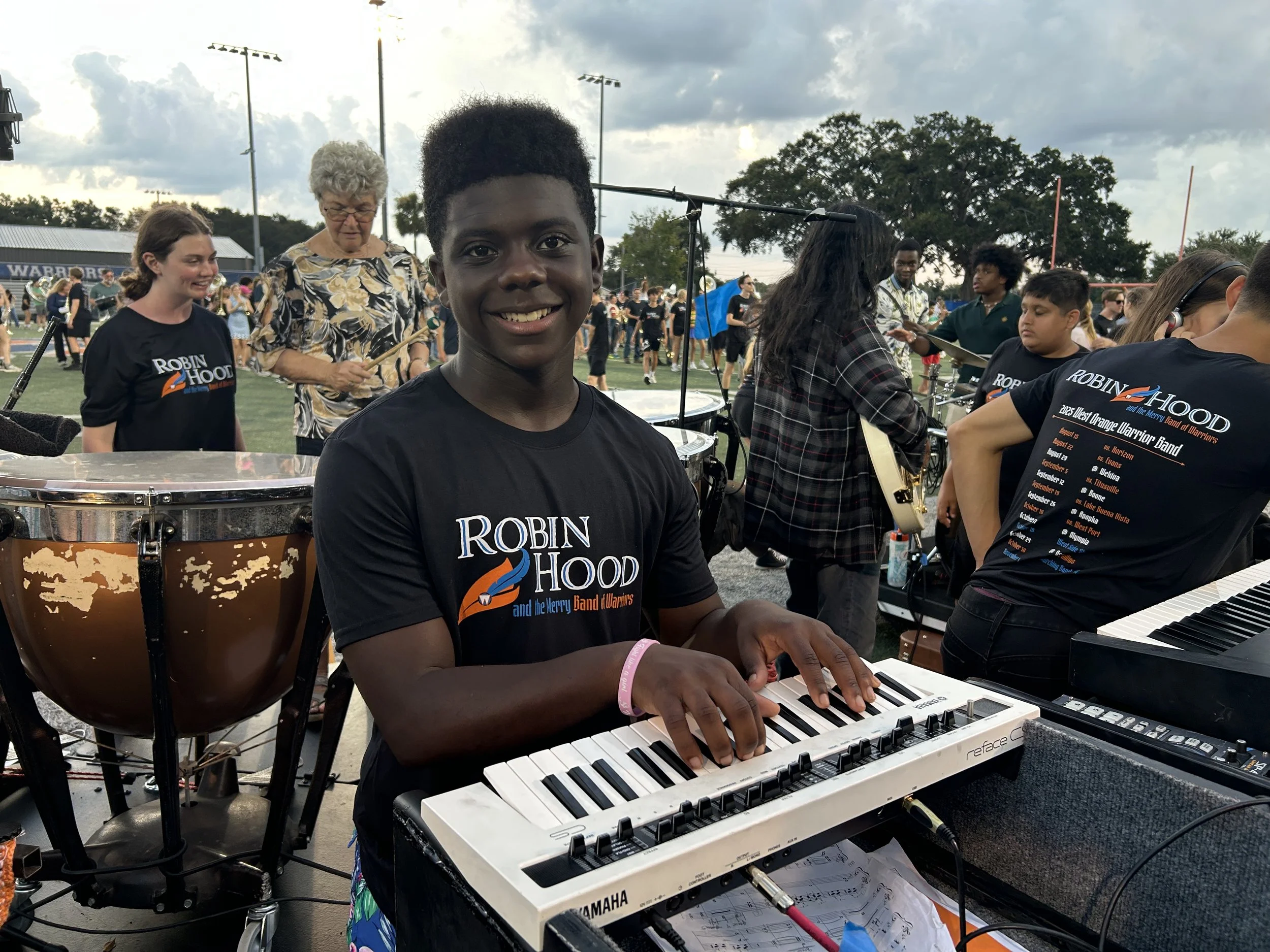 A young boy playing a white keyboard during an outdoor musical performance with a large group of people in the background.
