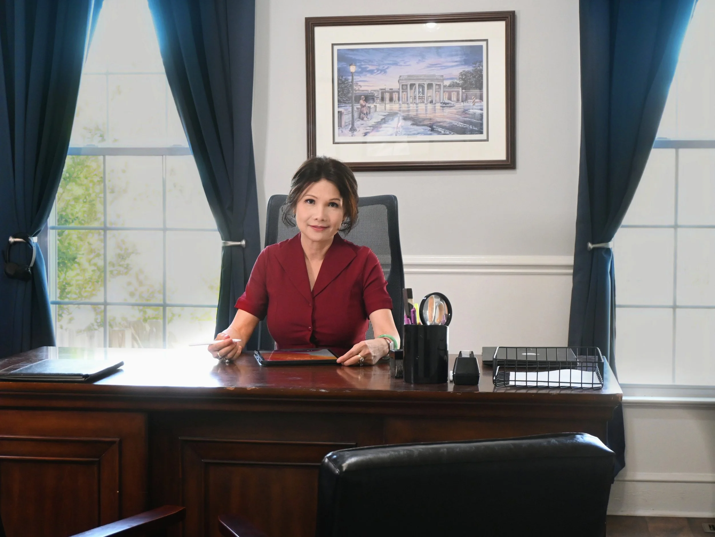 A financial analyst woman sitting at a large wooden desk in an office, with a tablet, office supplies, and a desktop organizer, in front of a window with blue curtains and a framed picture of a building on the wall.