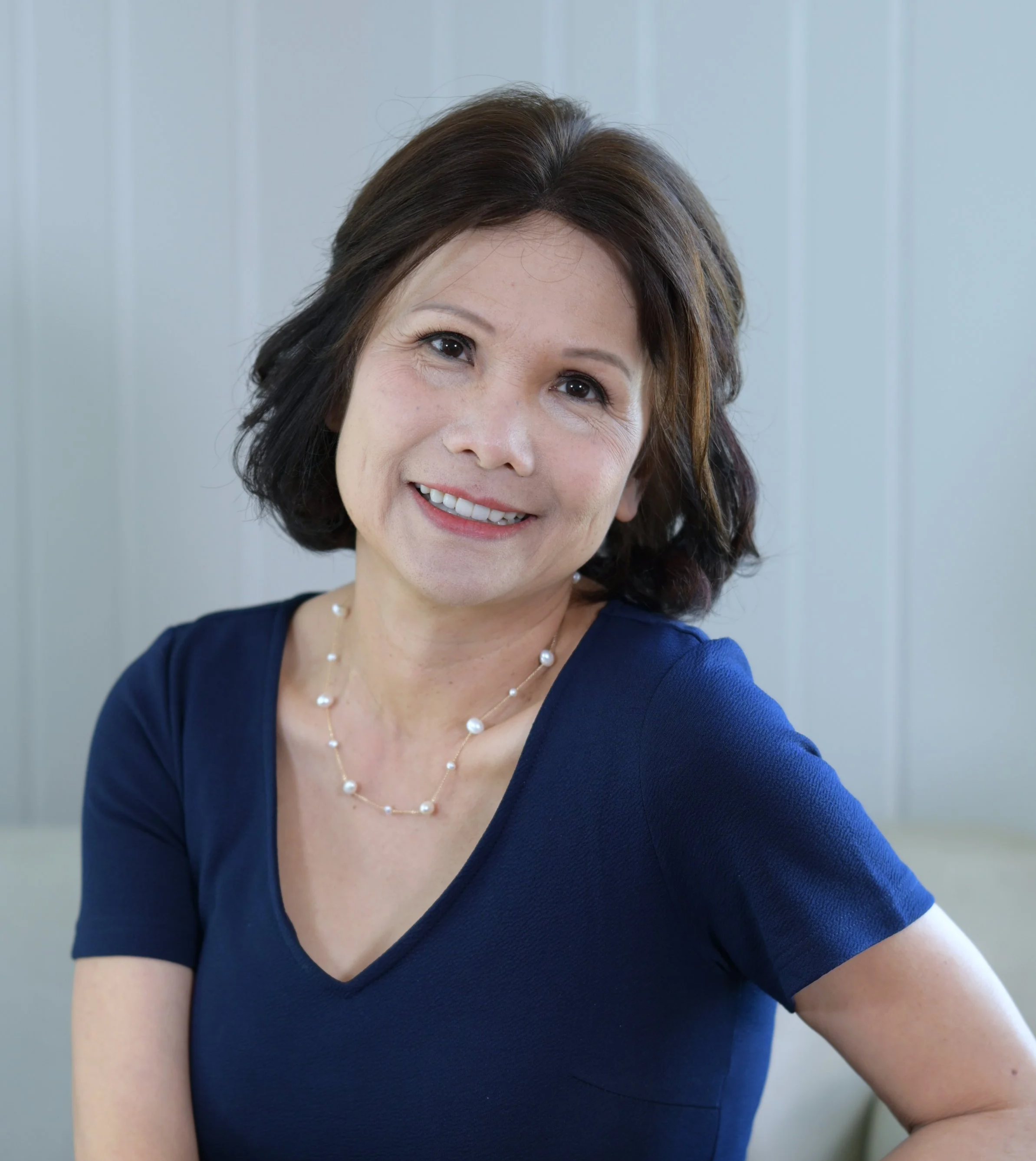 A woman with shoulder-length brown hair wearing a navy blue top and a pearl necklace, smiling while sitting against a light-colored panel background.