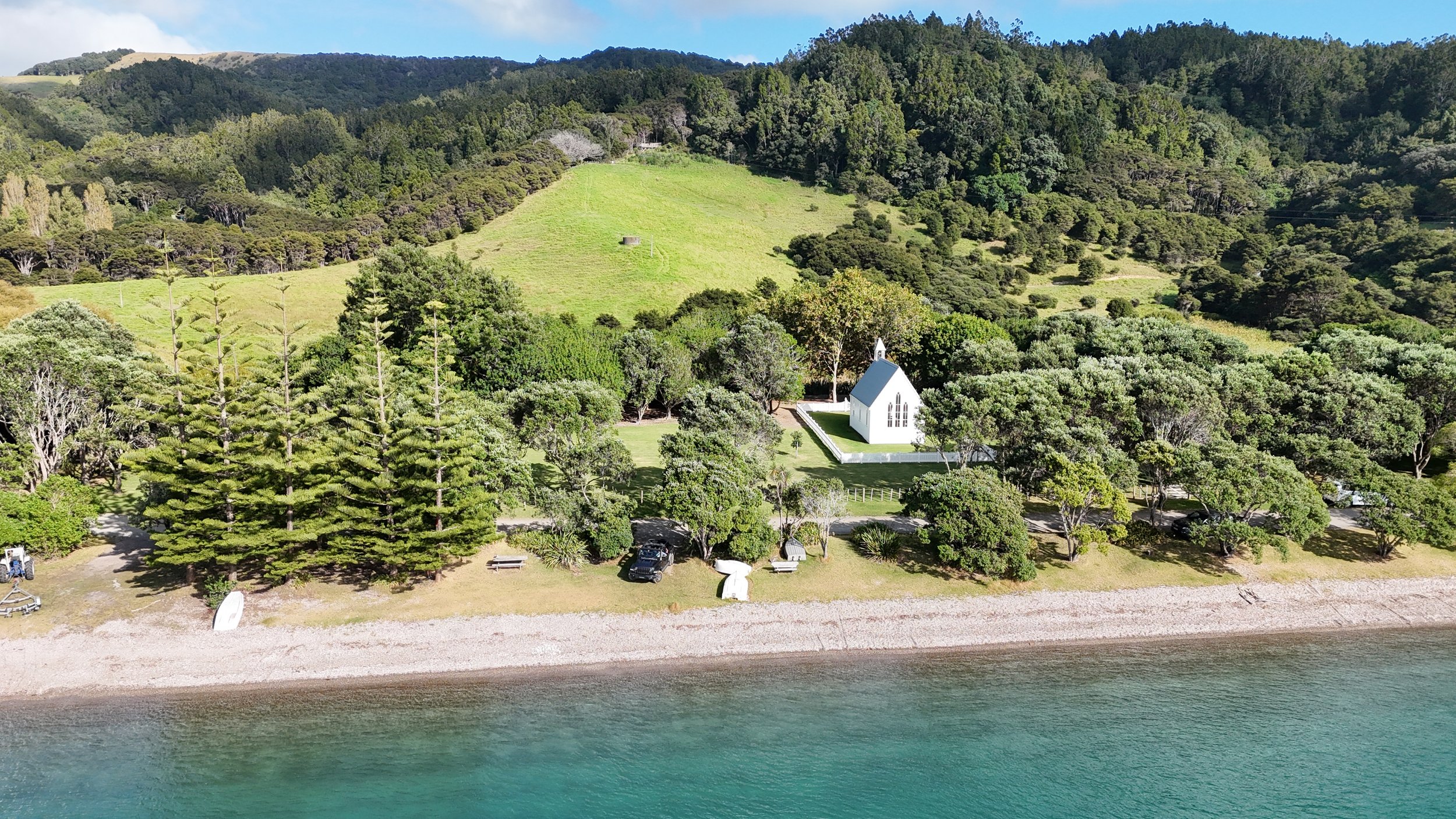 A small white church with a black roof situated among trees next to a gravel beach, with a grassy hillside covered in dense trees and a forested mountain in the background.
