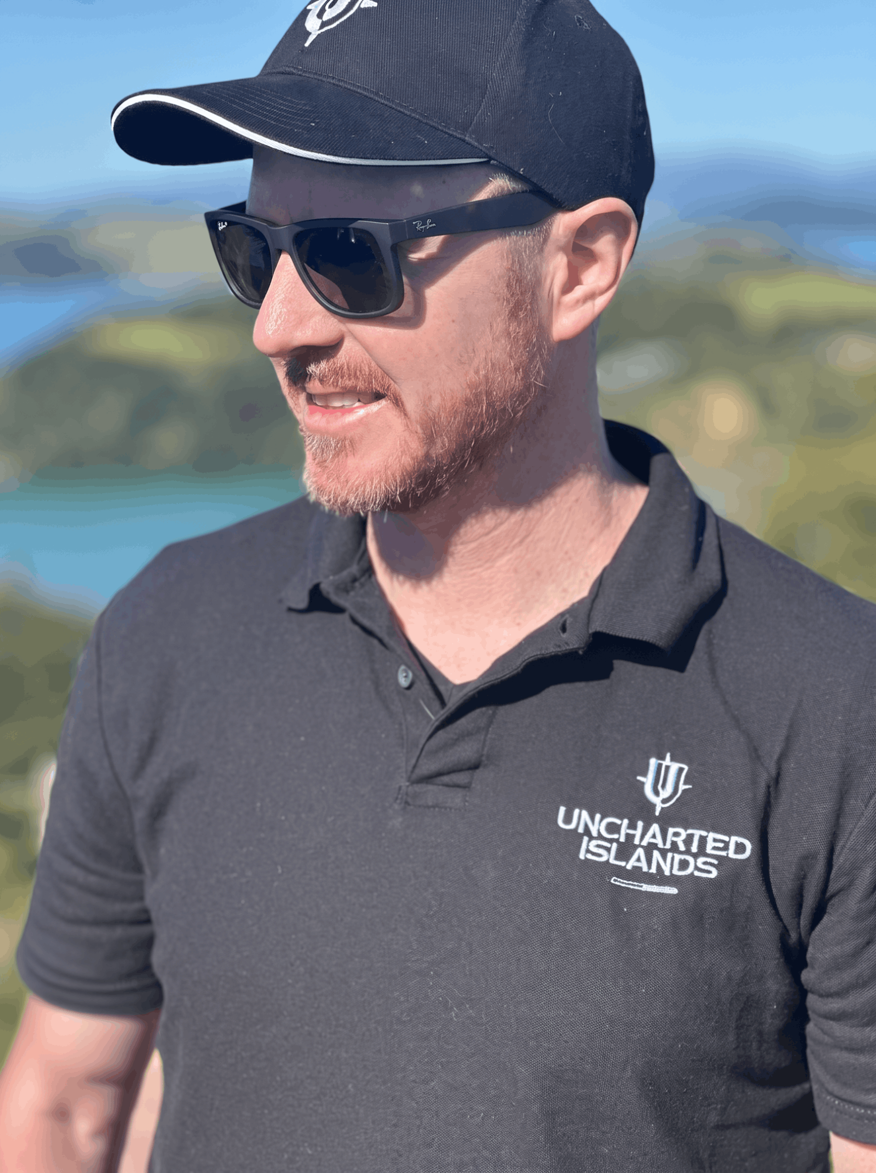 Close-up of a man wearing a black cap with a white logo, sunglasses, and a black polo shirt with the logo 'Uncharted Islands' and a cactus icon embroidered on the chest, standing outdoors with a blurred background of greenery and sky.