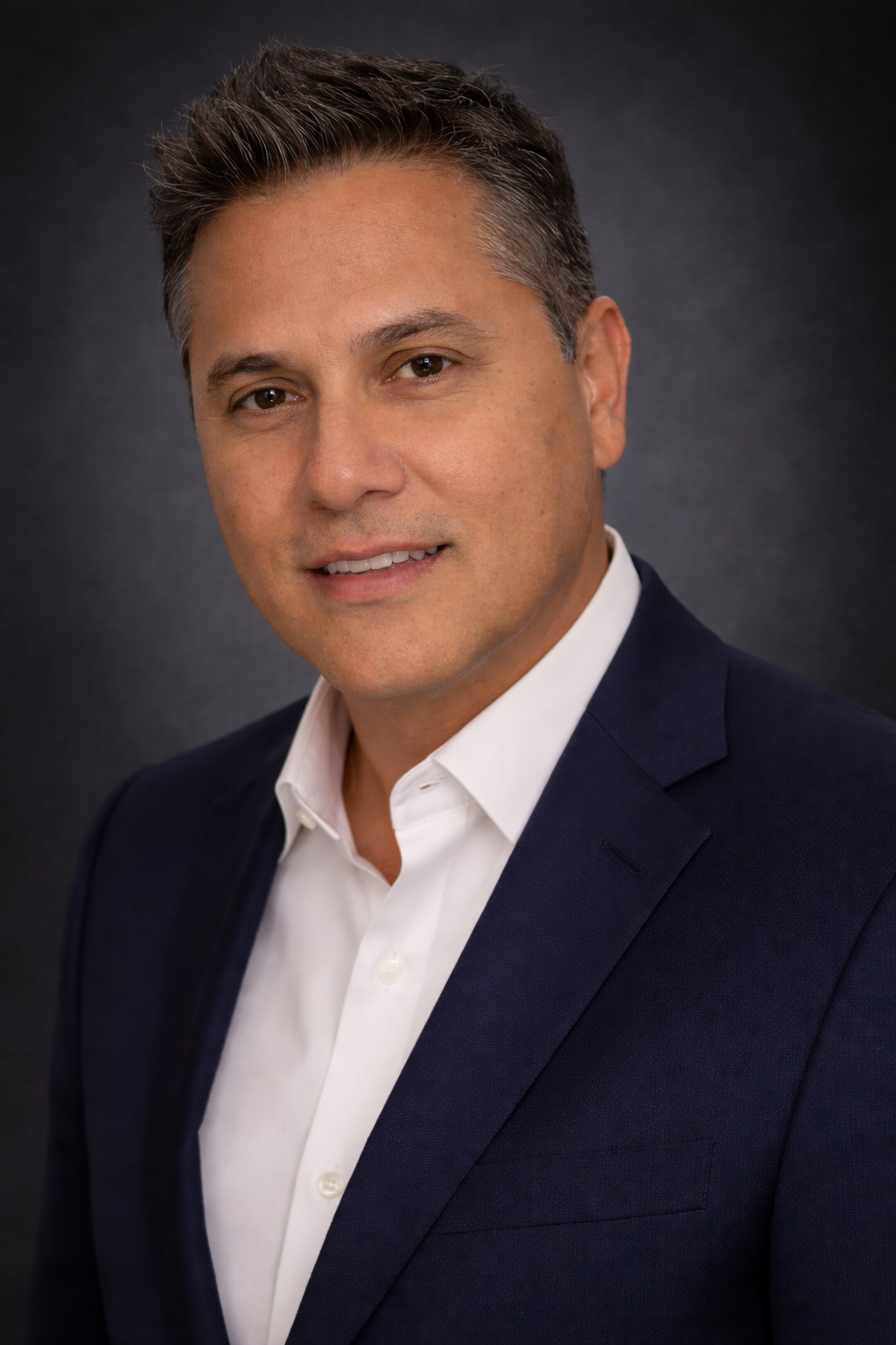 A professional headshot of Nicolas Miramontes with short, styled hair, wearing a dark suit jacket and a white dress shirt, smiling slightly against a dark, plain background.