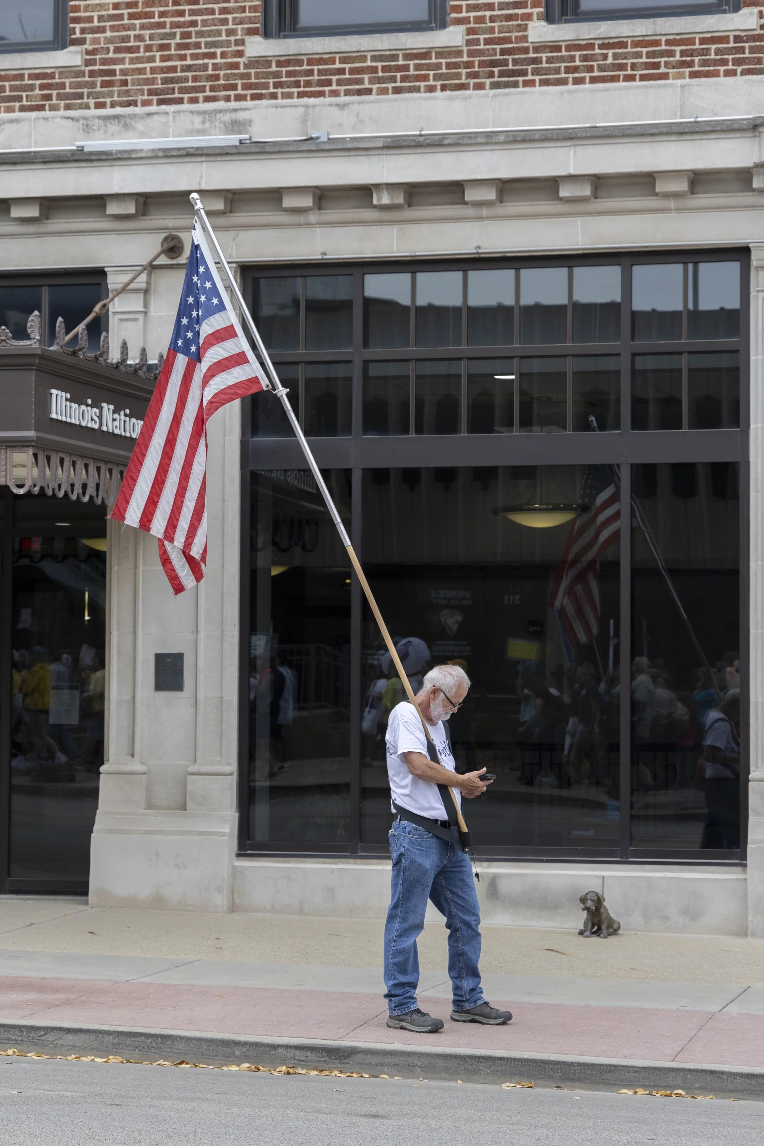 Bob Pointer looks at his phone after he places the large flag in its stand on his belt while the protest is marching towards the Old Capitol Building on the other side of the street at the No Kings protest Oct. 18, 2025 in front of Illinois National 