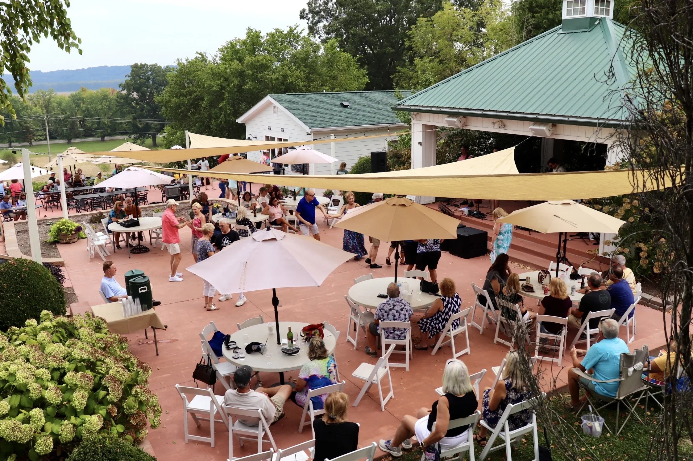 An outdoor event at a patio during daytime with people dancing, sitting at tables, and socializing, some under umbrellas and shade sails, with a house and green trees in the background.