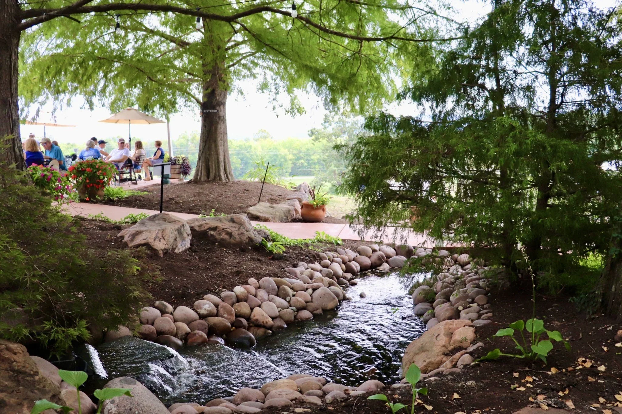 People sitting under umbrellas and trees in a garden or park area near a small stream with rocks and plants.