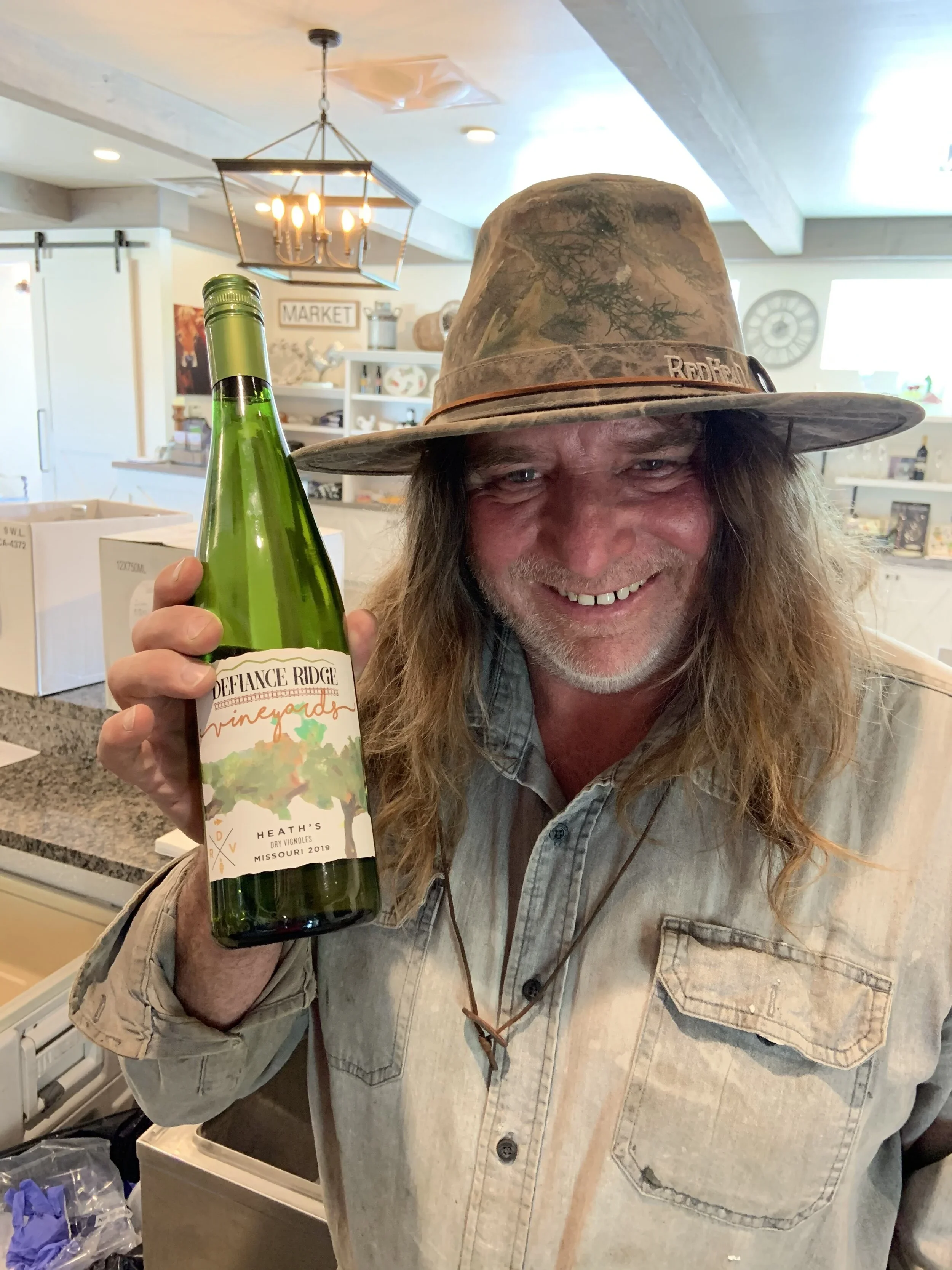 Smiling man with long hair and beard wearing a camouflage hat and light-colored button-up shirt, holding a green wine bottle labeled 'DeFlance Ridge Vineyards' in a kitchen.