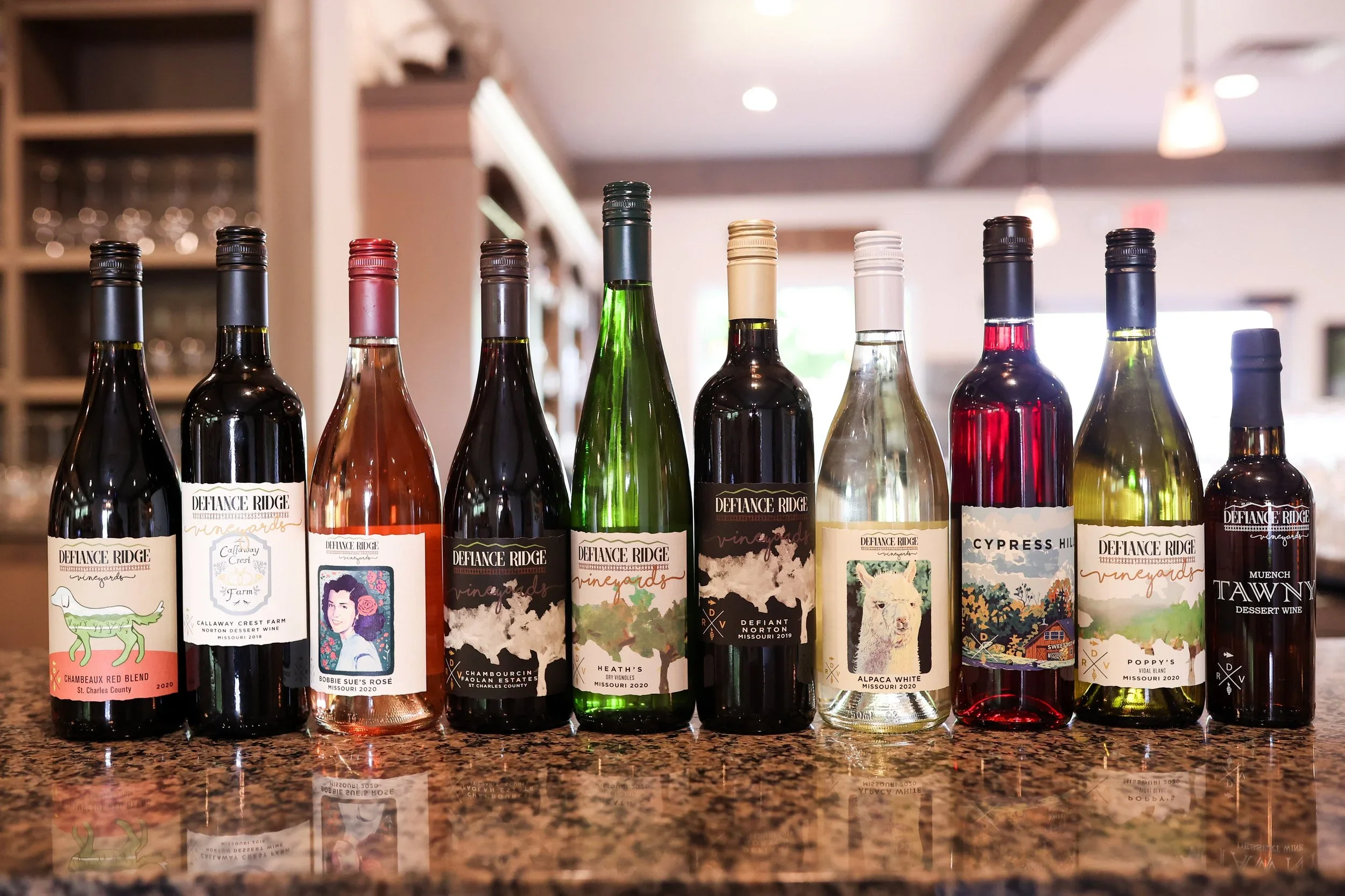 A row of nine different bottles of wine on a granite countertop in a home kitchen, with blurred background of kitchen cabinets and lighting.