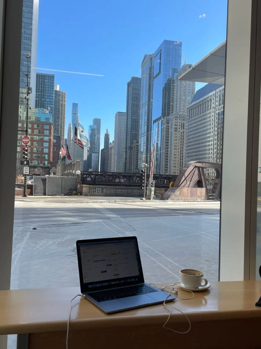 A cafe window view of a cityscape with tall modern buildings, flags, and clear blue sky, with a laptop and coffee on the table inside the cafe.