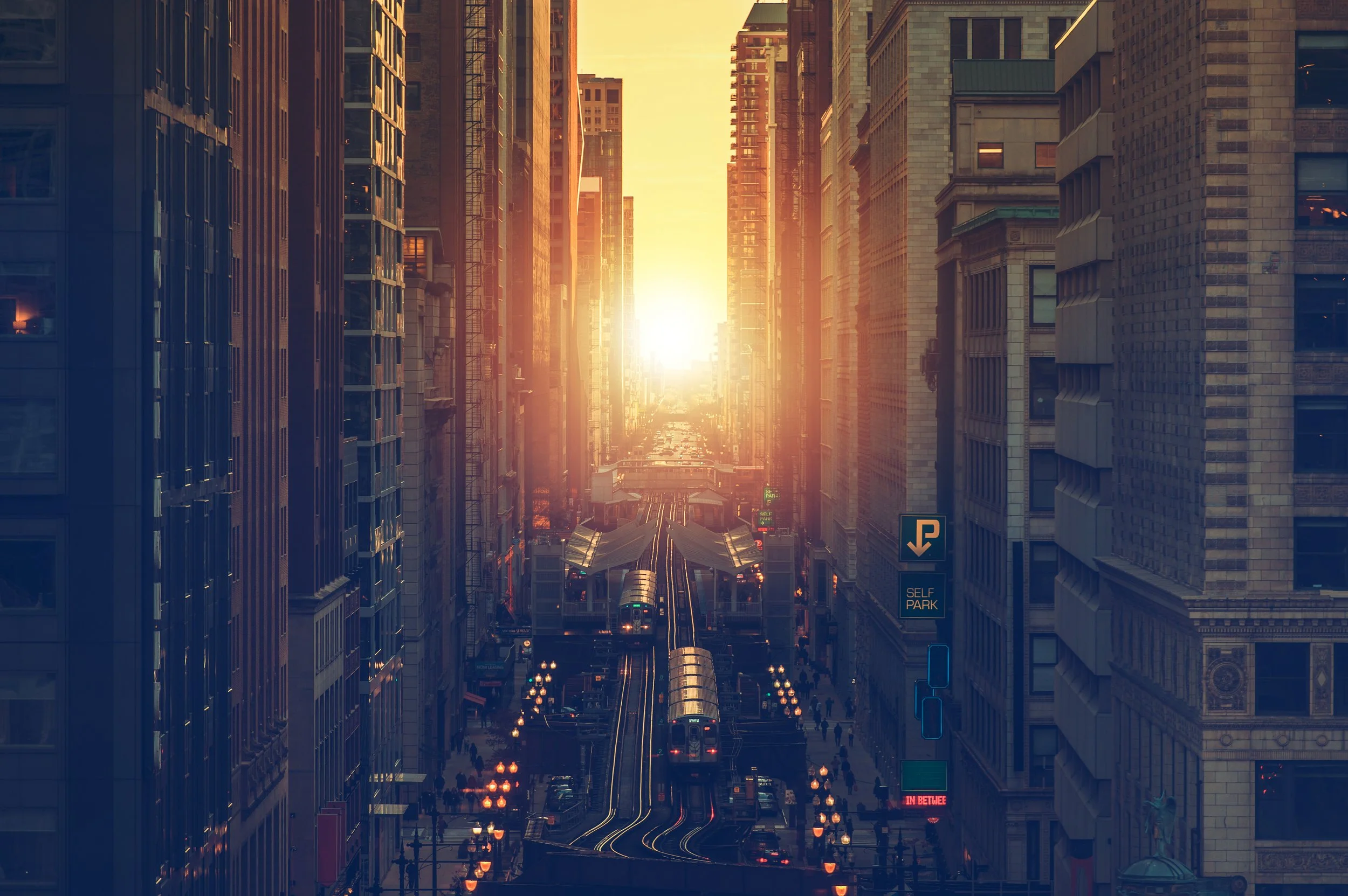 City street at sunset with tall buildings on both sides, a railway with trains, and streetlights illuminating the area.