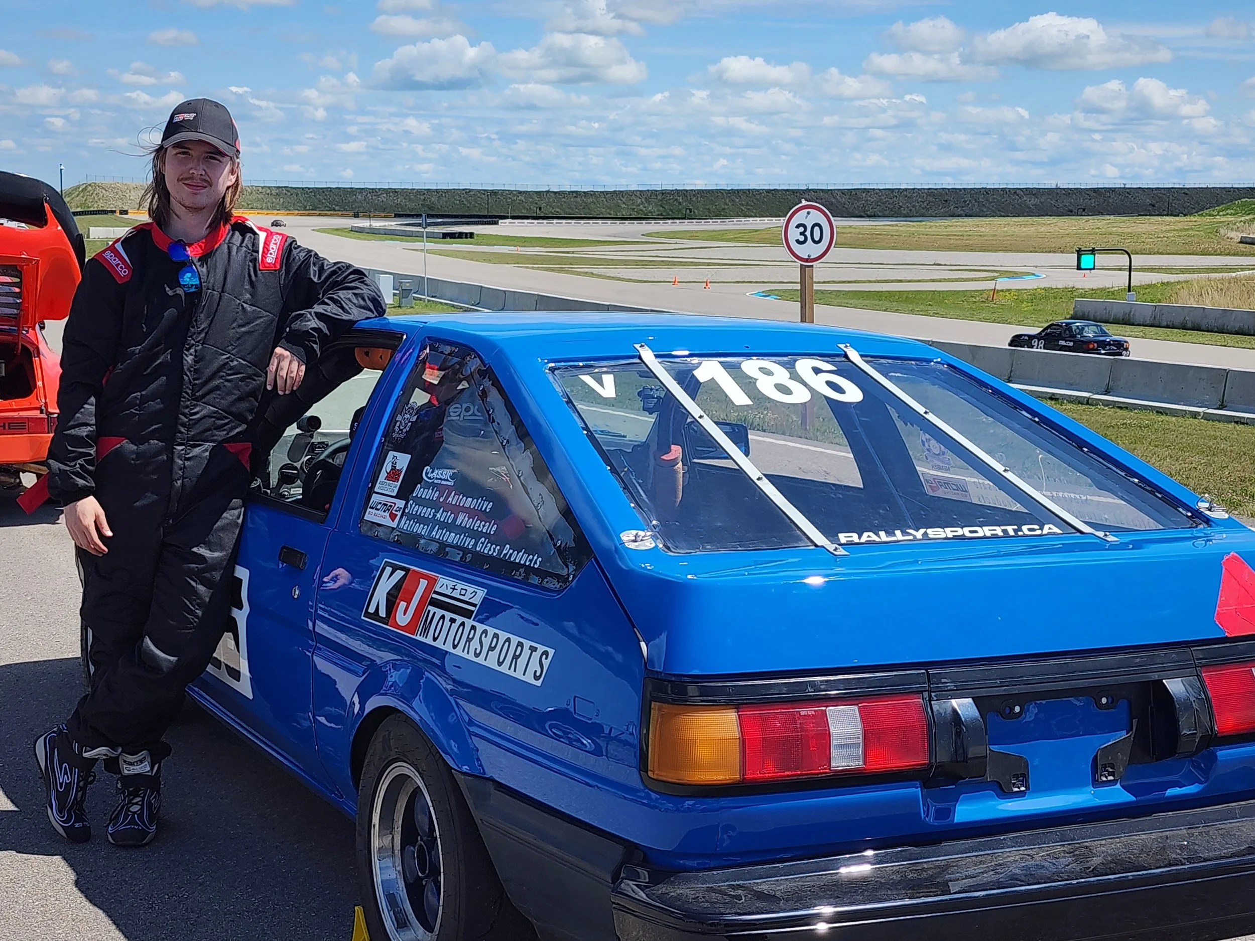 Young man in racing suit and cap standing next to blue rally car at racetrack on a sunny day.
