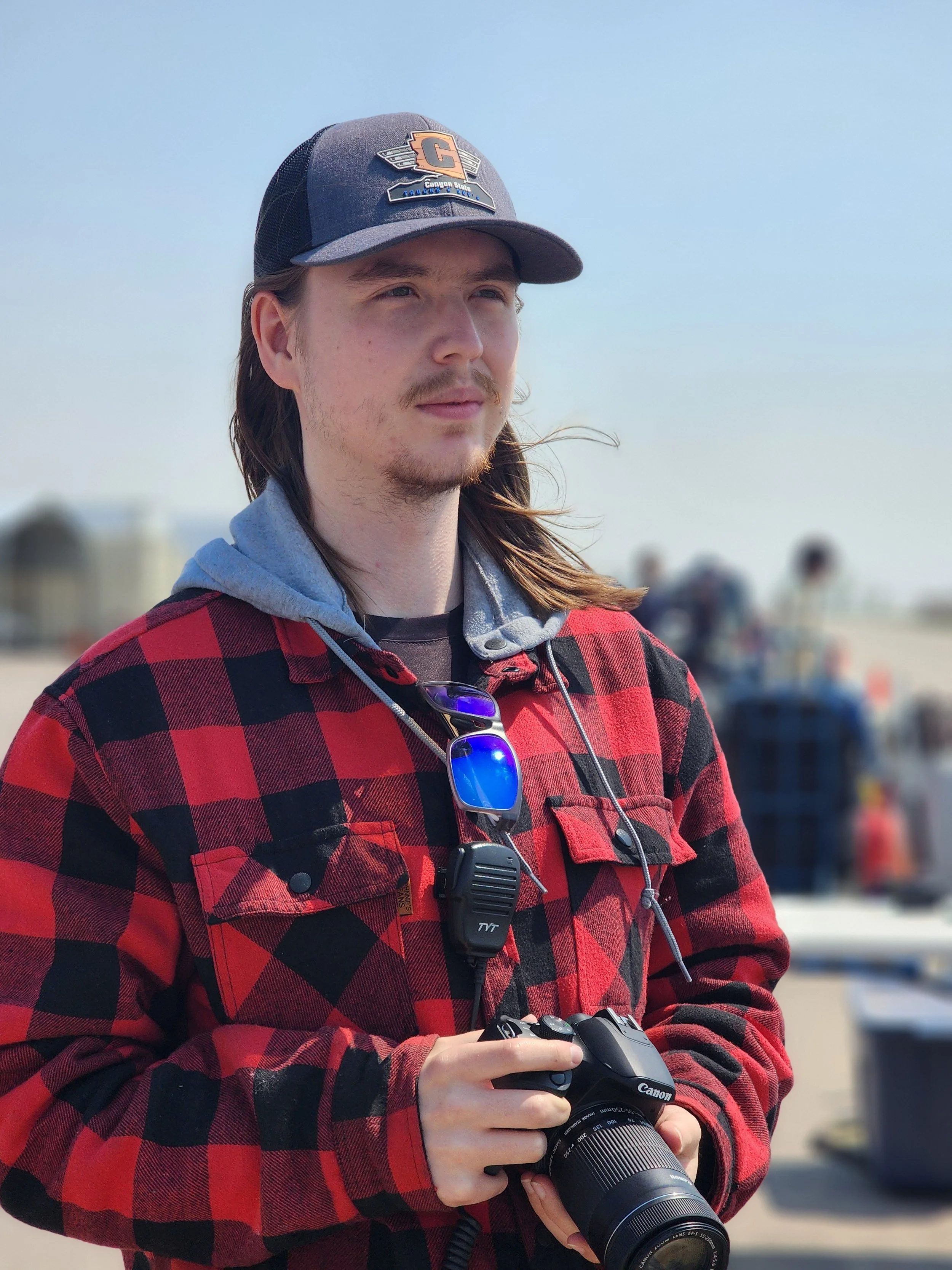 Kade Stevens a baseball cap, a red and black plaid jacket, holding a camera. Kade Stevens is at an Alberta Race Car Association event photographing and capturing social media content.