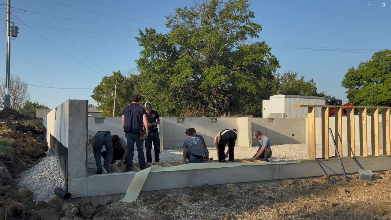 Six people working on a construction site with concrete walls and a wooden frame, surrounded by dirt and gravel, under a clear sky with trees in the background.