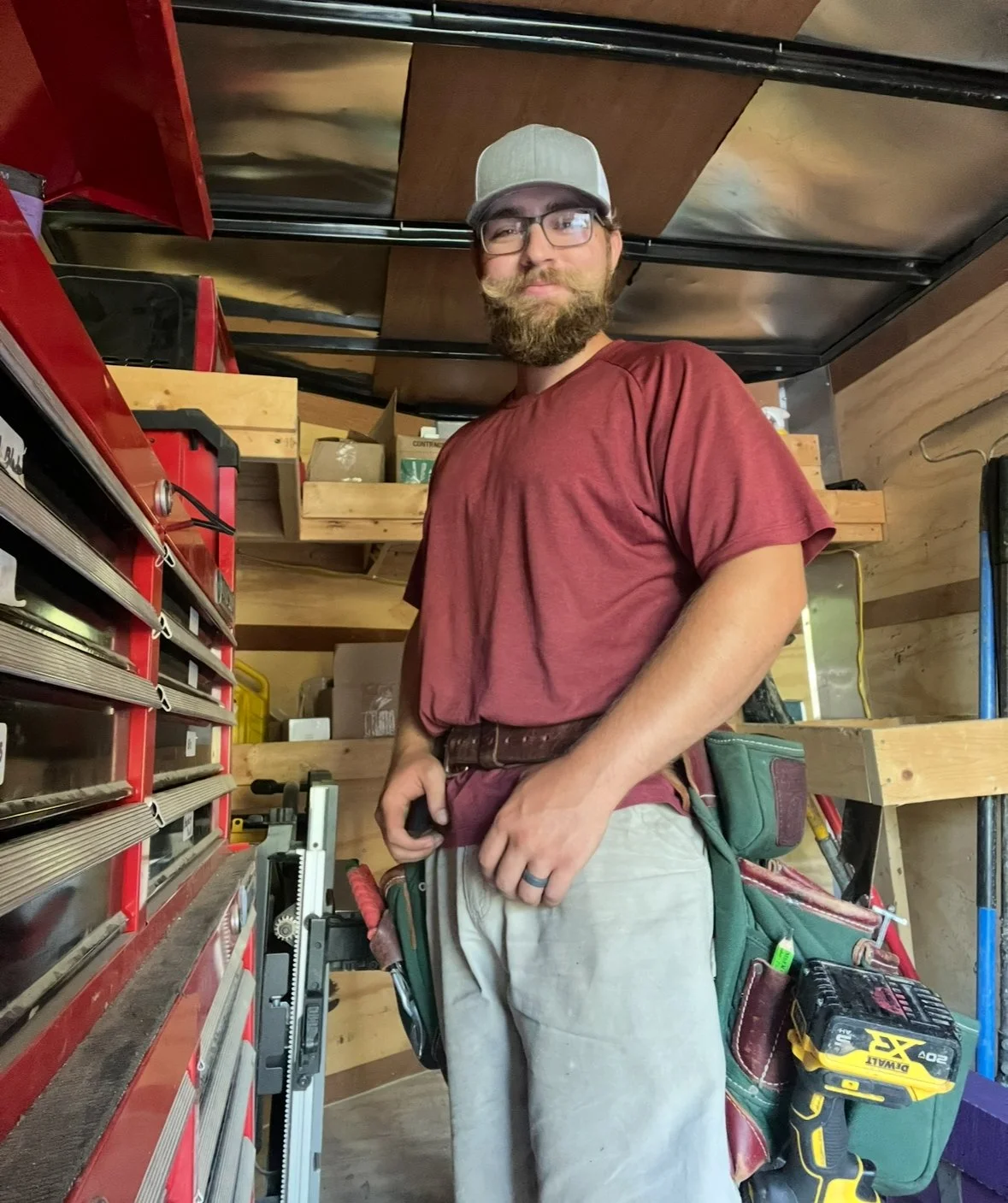 Man with glasses, beard, and mustache, wearing a gray cap, maroon t-shirt, and beige work pants, standing in a workshop with a tool belt containing a yellow and black drill, among shelves and storage containers.