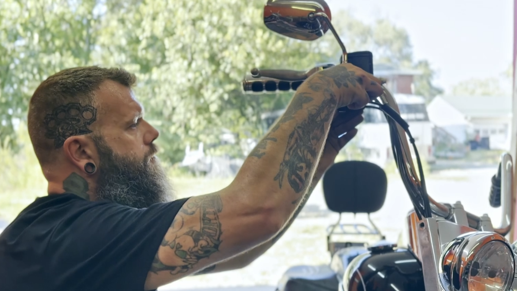 A man with tattoos, a beard, and earrings adjusting the handlebar of a motorcycle inside a garage or workshop with a window showing trees and houses outside.