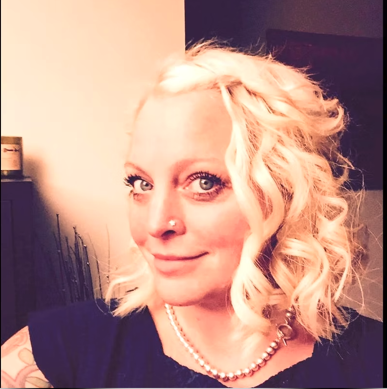 A woman with blonde, curly hair, blue eyes, and a nose piercing, smiling for a selfie indoors, wearing a pearl necklace and a dark top.