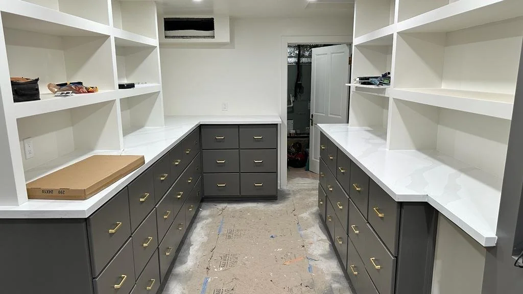 Empty kitchen with white open shelving, dark gray cabinets with brass handles, and white marble countertops, in a room under renovation.