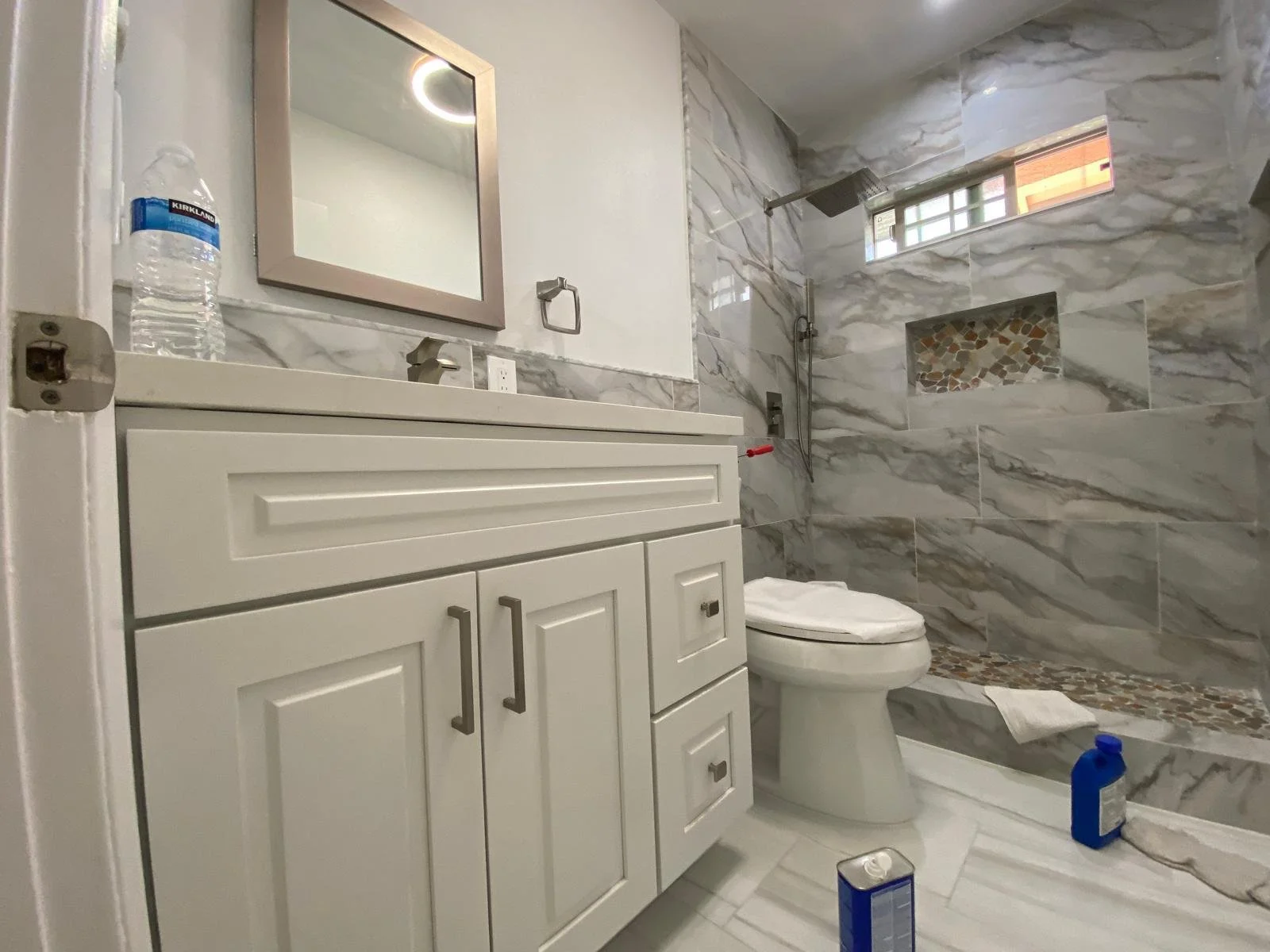 Modern bathroom with marble tiles, a white vanity with silver handles, a framed mirror, a bottle of water on the counter, a toilet, a showerhead, a small window, and cleaning supplies on the floor.