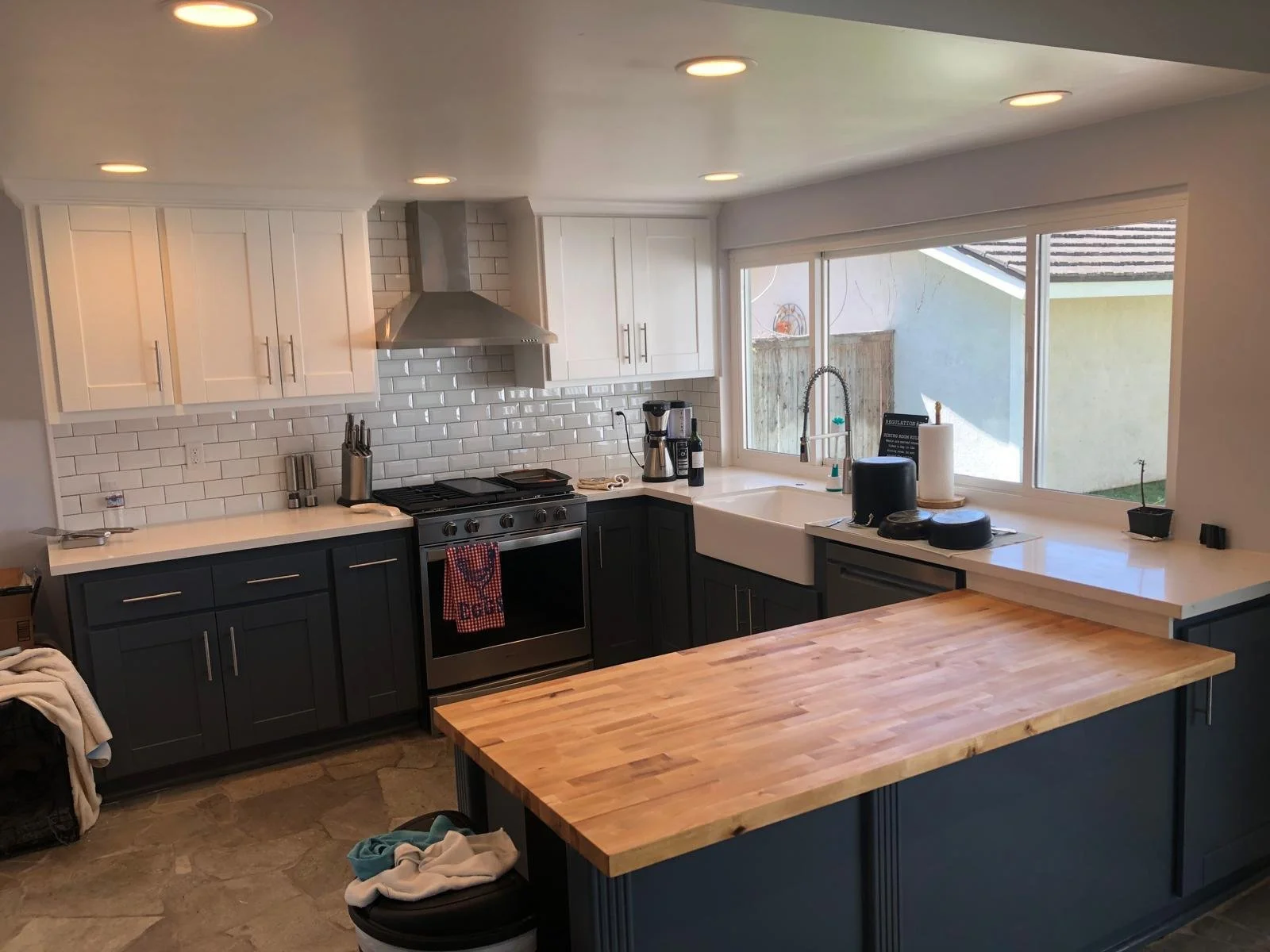 Modern kitchen with white upper cabinets, dark lower cabinets, a white subway tile backsplash, stainless steel appliances, a large farmhouse sink, a wooden kitchen island, and large windows over the sink letting in natural light.