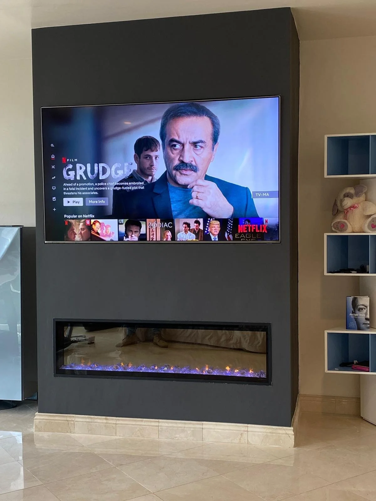 Living room with a wall-mounted flat-screen TV displaying a movie called 'Grudge'. Below the TV is a modern electric fireplace with purple and orange flames. To the right, there is a white bookshelf with a teddy bear, a book, and other objects.