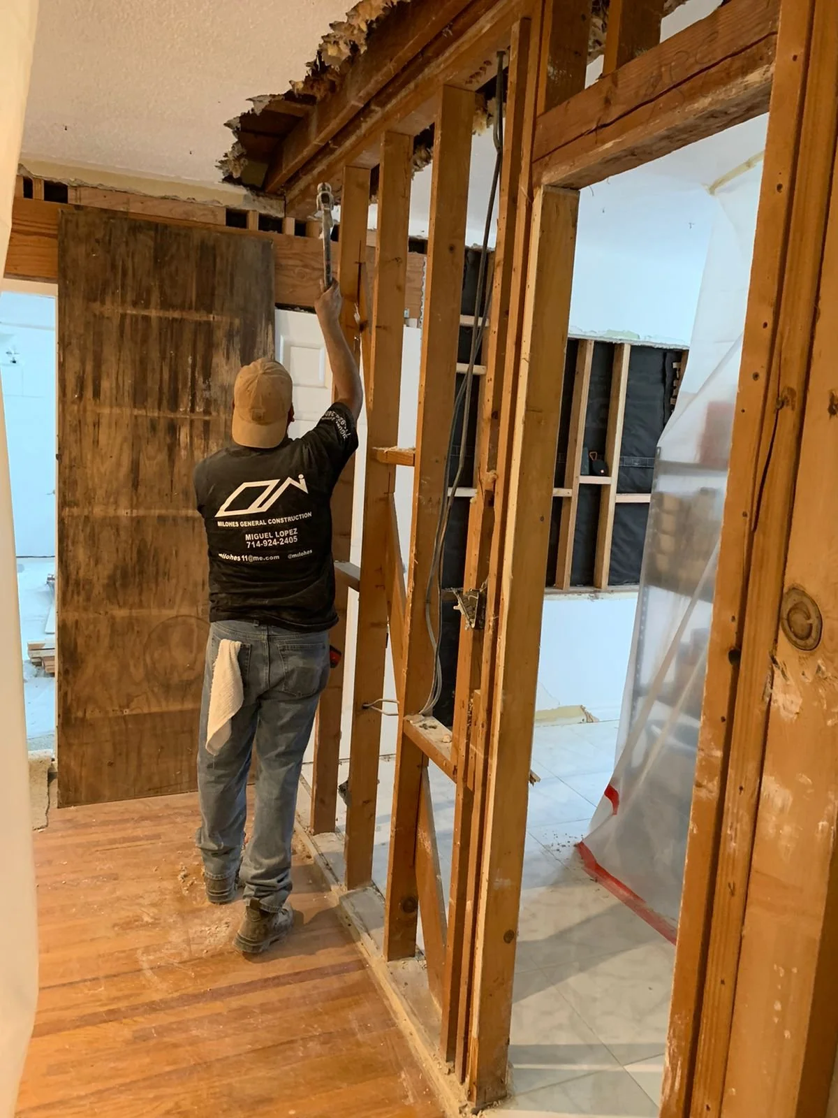 A construction worker wearing jeans and a black shirt with the logo for Milones General Construction, applying white paint or primer to the top of a wooden wall frame in a room under renovation. The room has exposed wooden studs, some drywall, and a partially completed doorway leading to a tiled floor area.