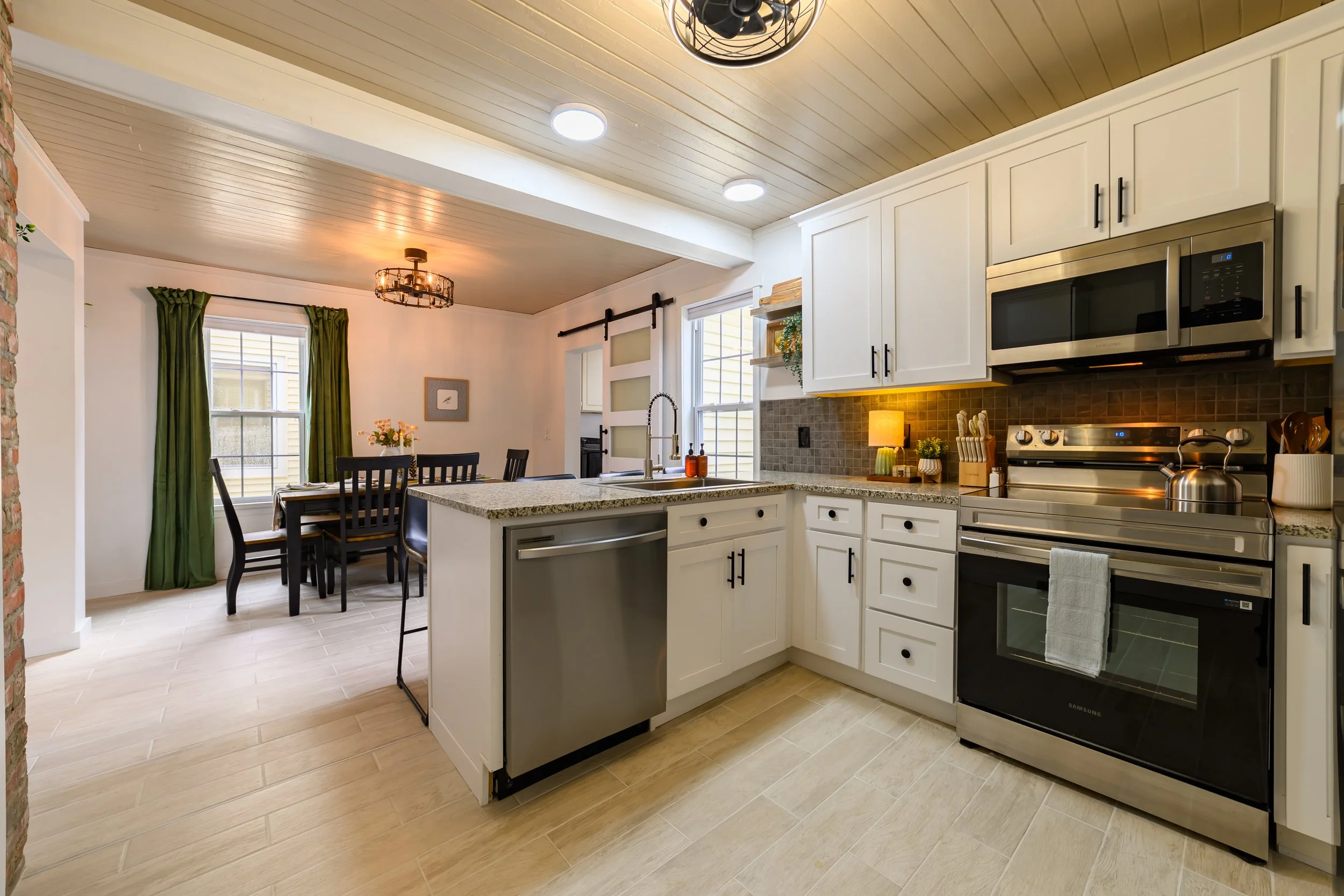 Modern kitchen with white cabinets, stainless steel appliances, granite countertops, and a dining area with a dark wood table and chairs, illuminated by ceiling lights and a small table lamp, with forest green curtains.