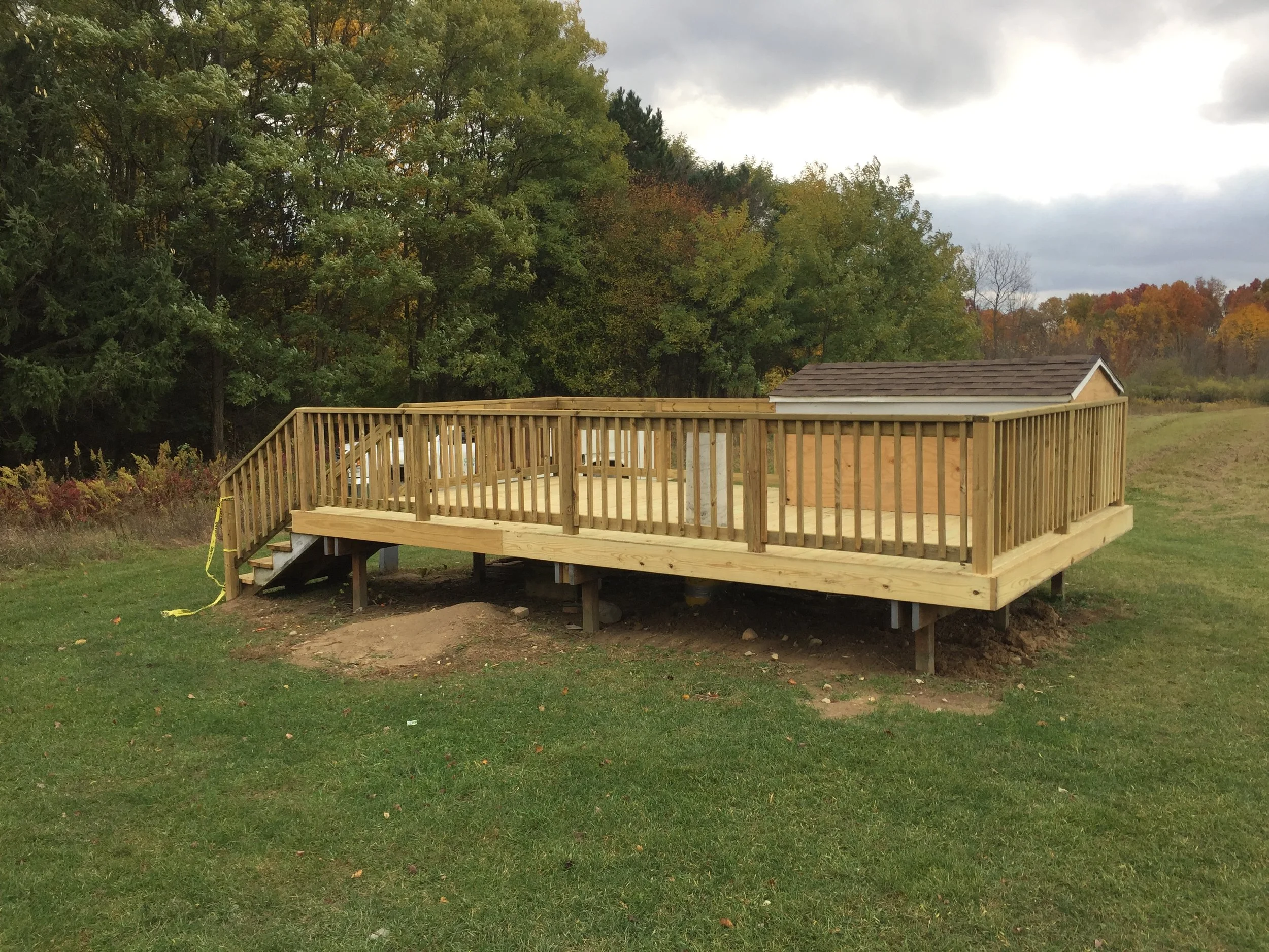 Recently built wooden deck with railing on a grassy backyard, with a small shed and trees with autumn foliage in the background.