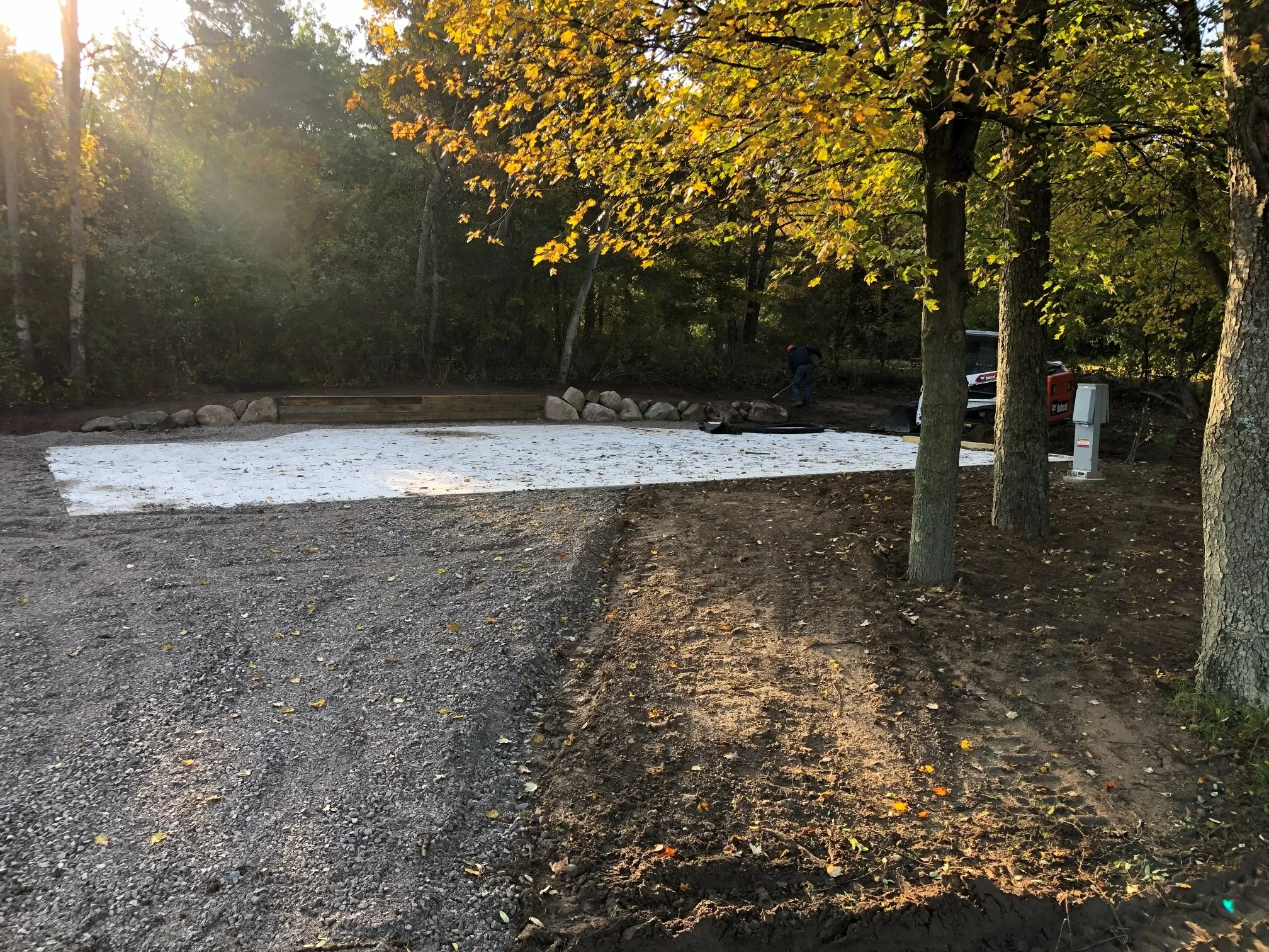 A construction site where a concrete pad is being prepared, with some workers and a car in the background, surrounded by trees.