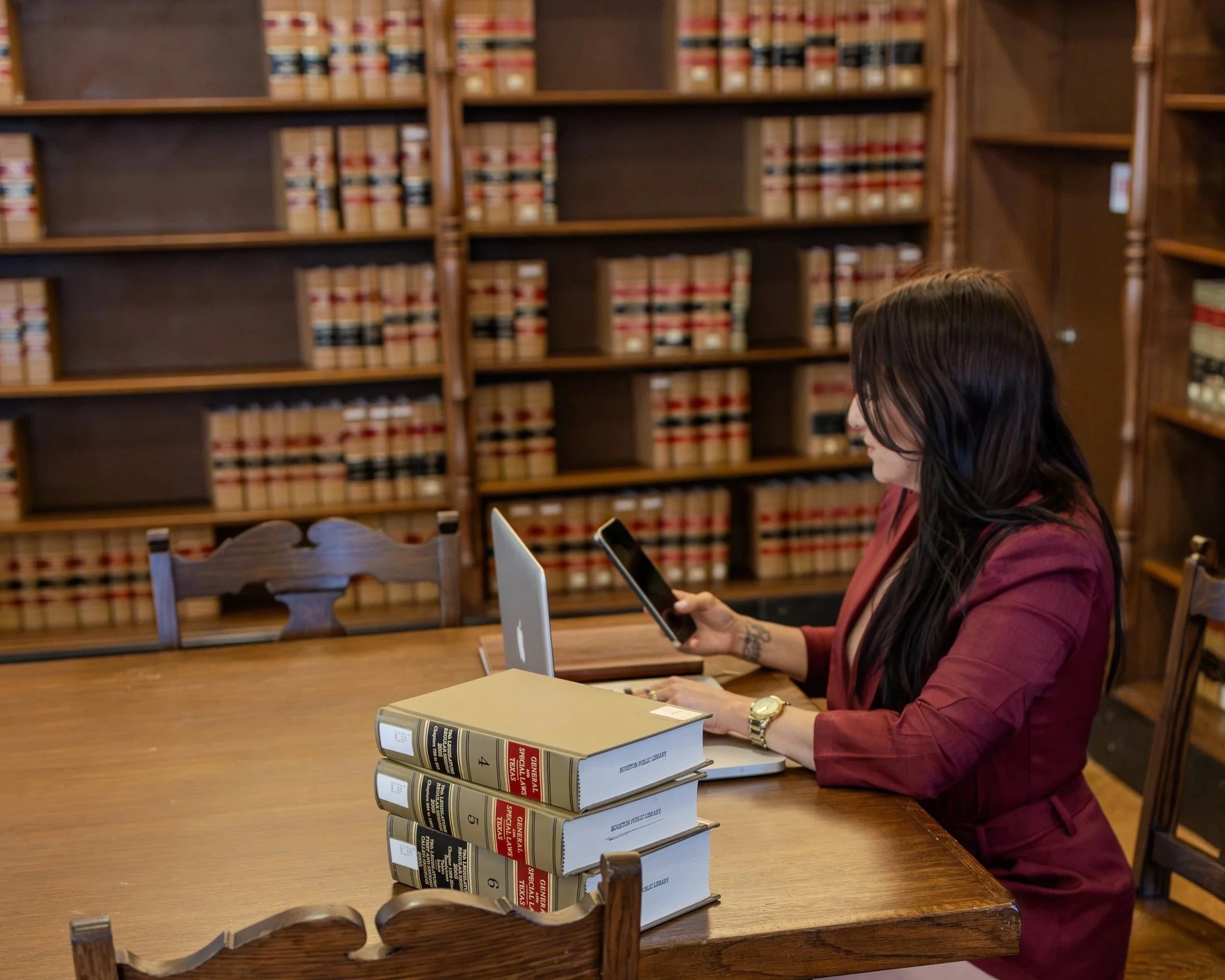 Woman in a maroon blazer sitting at a wooden table in a library, looking at her phone, with a stack of law books on the table and shelves of legal books in the background.