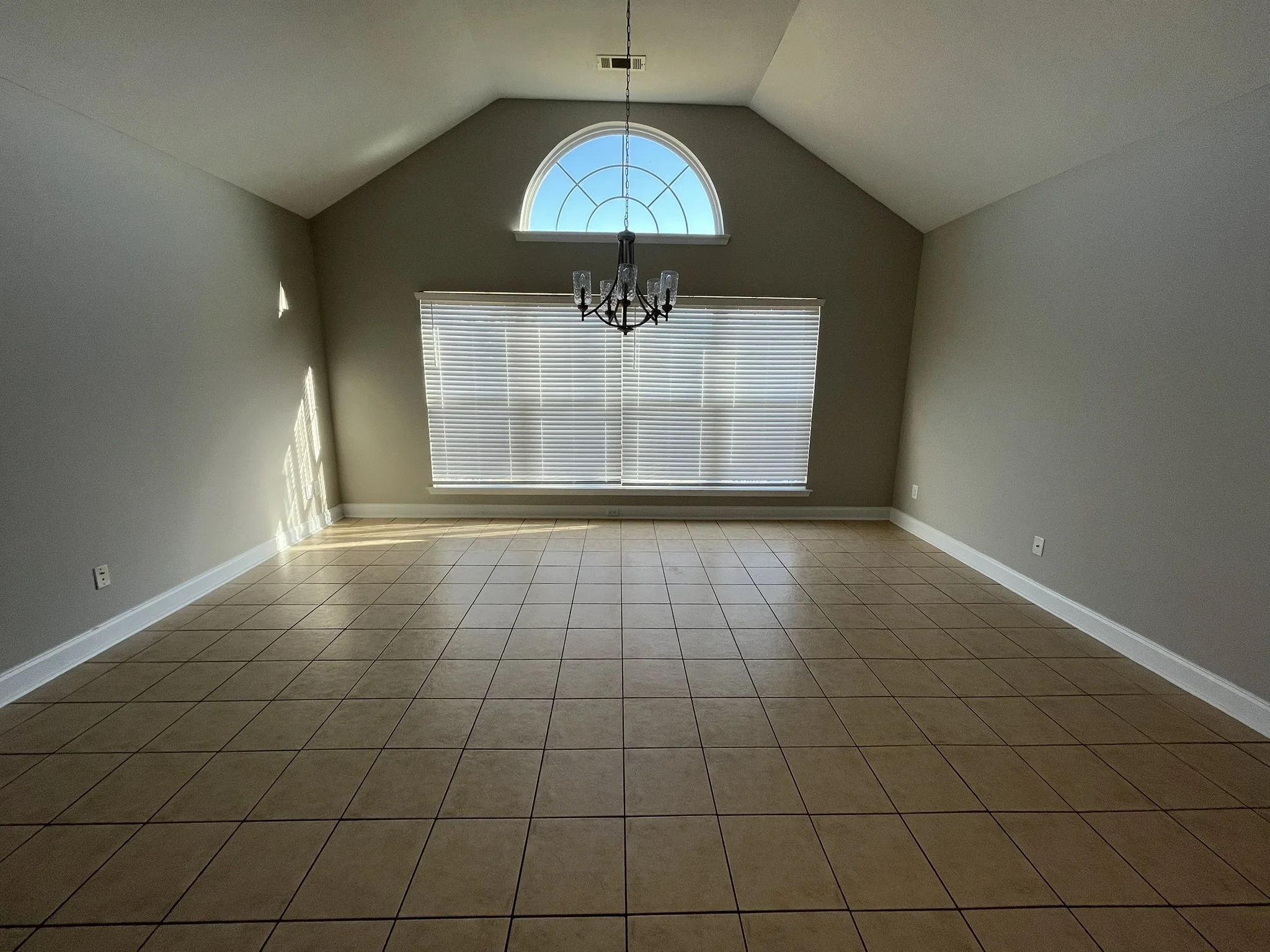 Before Virtual Staging. Empty living room with beige tiled floor, light gray walls, a large window with blinds, a semi-circular window above, and a chandelier hanging from the ceiling.