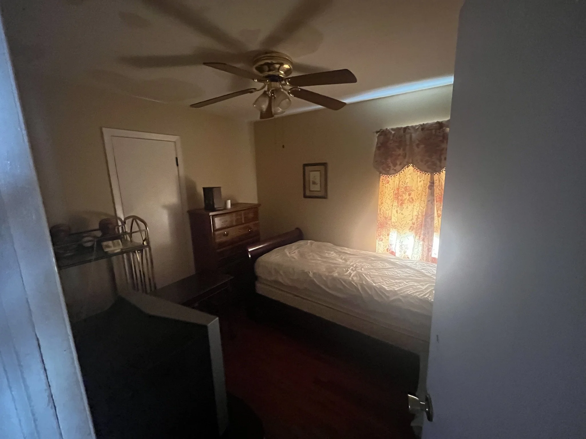 A dimly lit bedroom with a ceiling fan, a wooden bed frame with a mattress, a wooden dresser, a small framed picture on the wall, and a window with floral curtains.