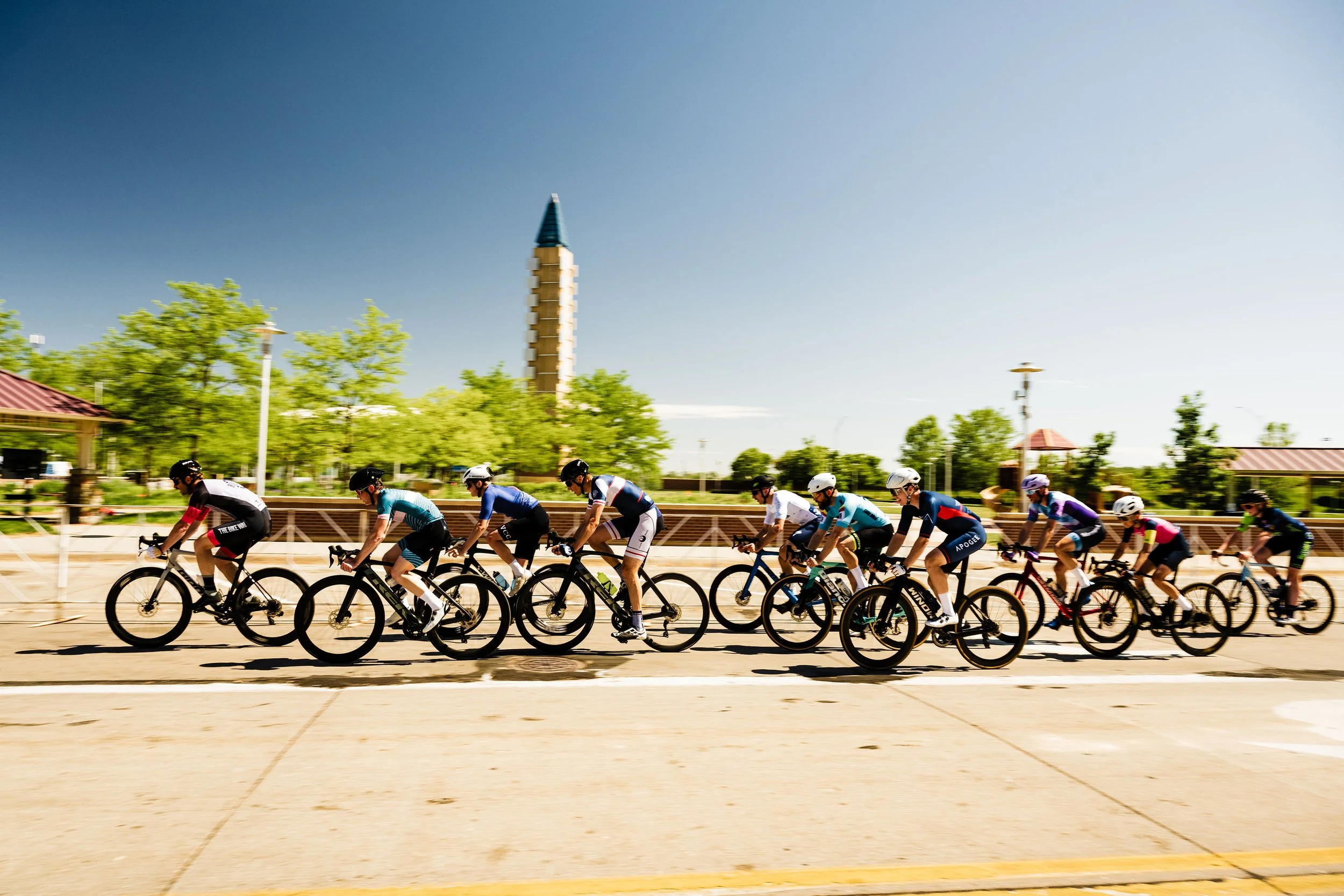 A group of bikers, racing in a criterium. They are in Stinson park, where you see a tower sculpture and  trees in the background.