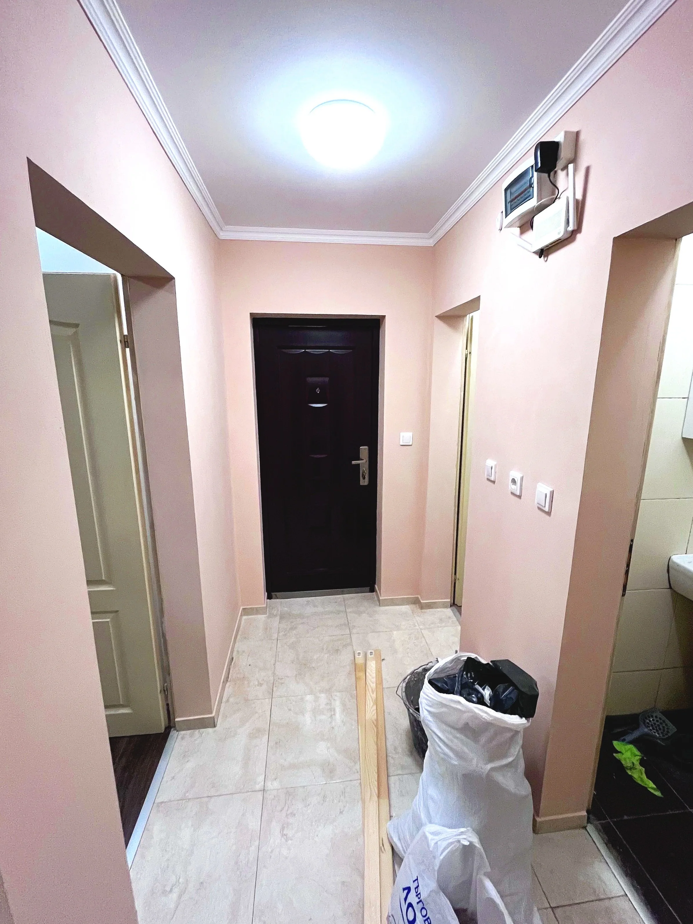 Hallway with a black door at the end, beige walls, white ceiling with crown molding, and tiled floor. There is a long wooden plank on the floor, a white garbage bag, a black garbage bag, and some tools and materials in the area.