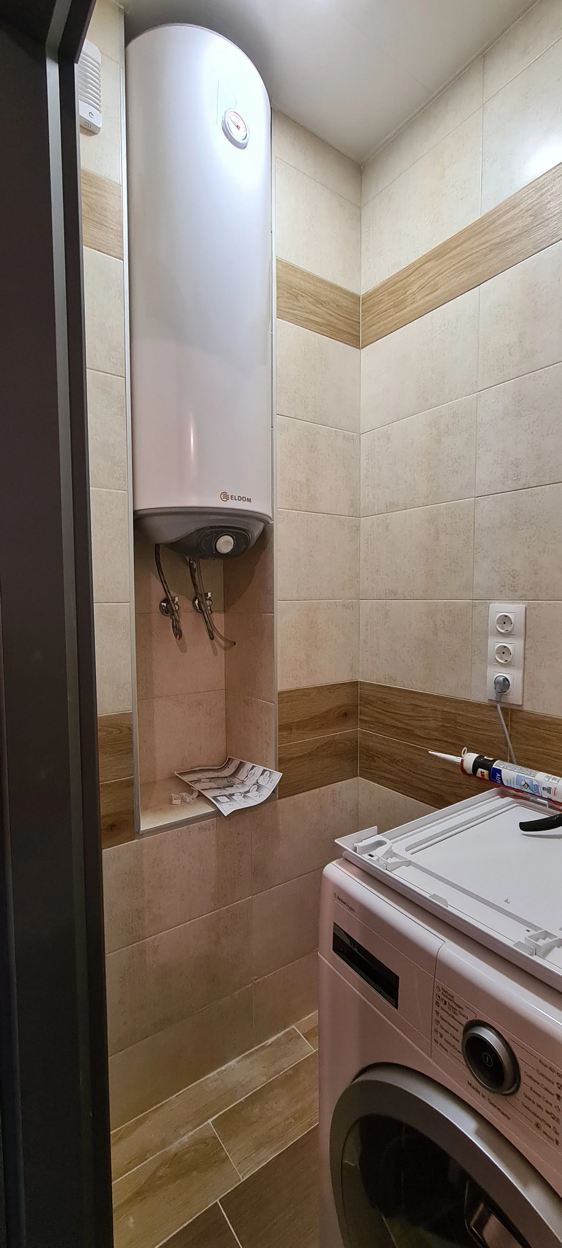 A laundry room with an electric water heater mounted on the wall, a washing machine, and an electrical outlet with plugs. There are some tools and documents on the shelf beneath the water heater.