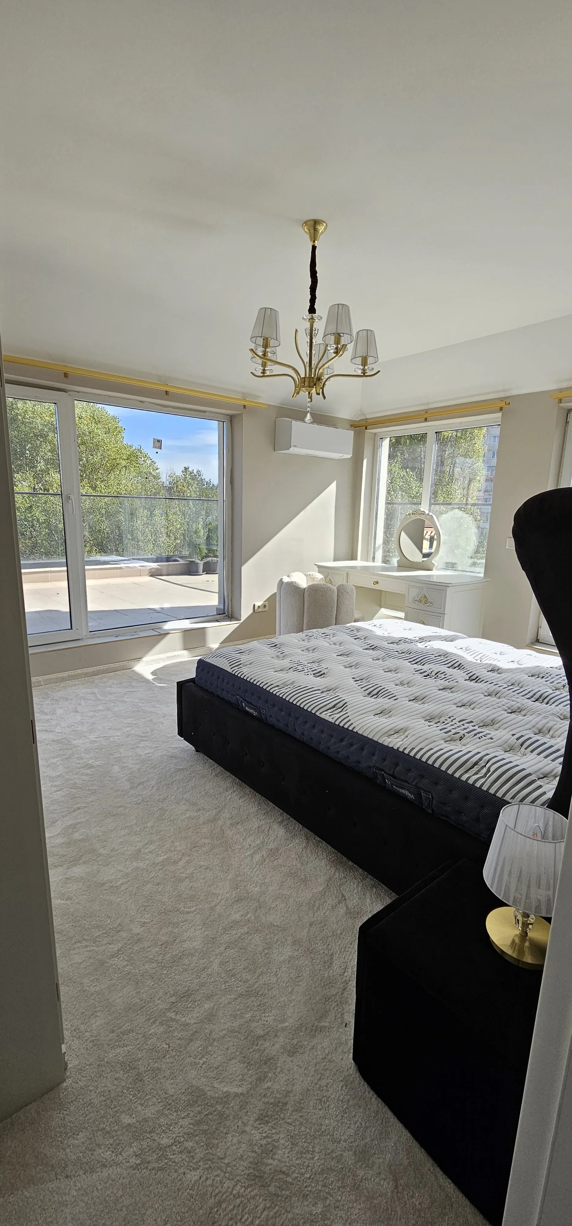 Bright bedroom with large windows, a white dresser with a mirror, a black bed frame with a striped mattress, a cream-colored armchair, and a chandelier hanging from the ceiling.