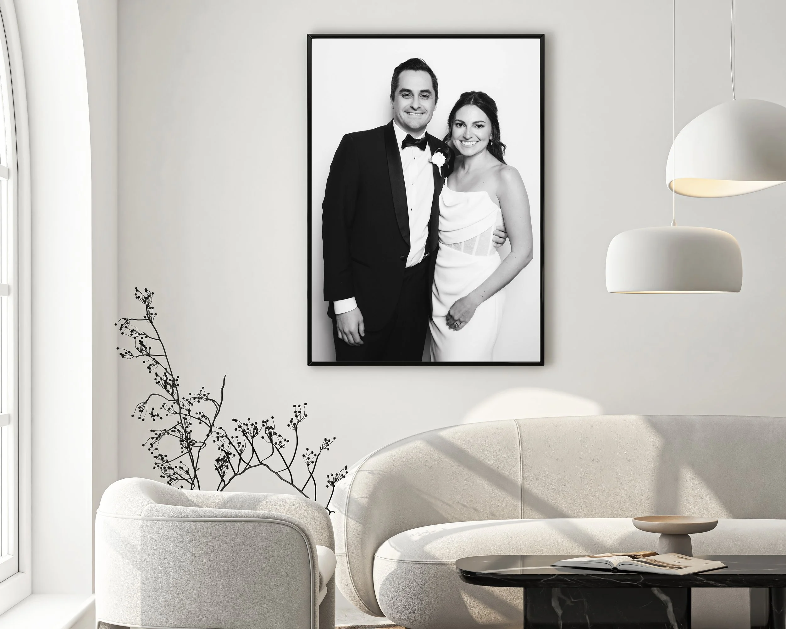 Modern living room with white curved sofas, a ceiling lamp, a small black table with a magazine, a framed black-and-white photograph of a smiling couple in wedding attire, and a decorative branch with black berries near the window.