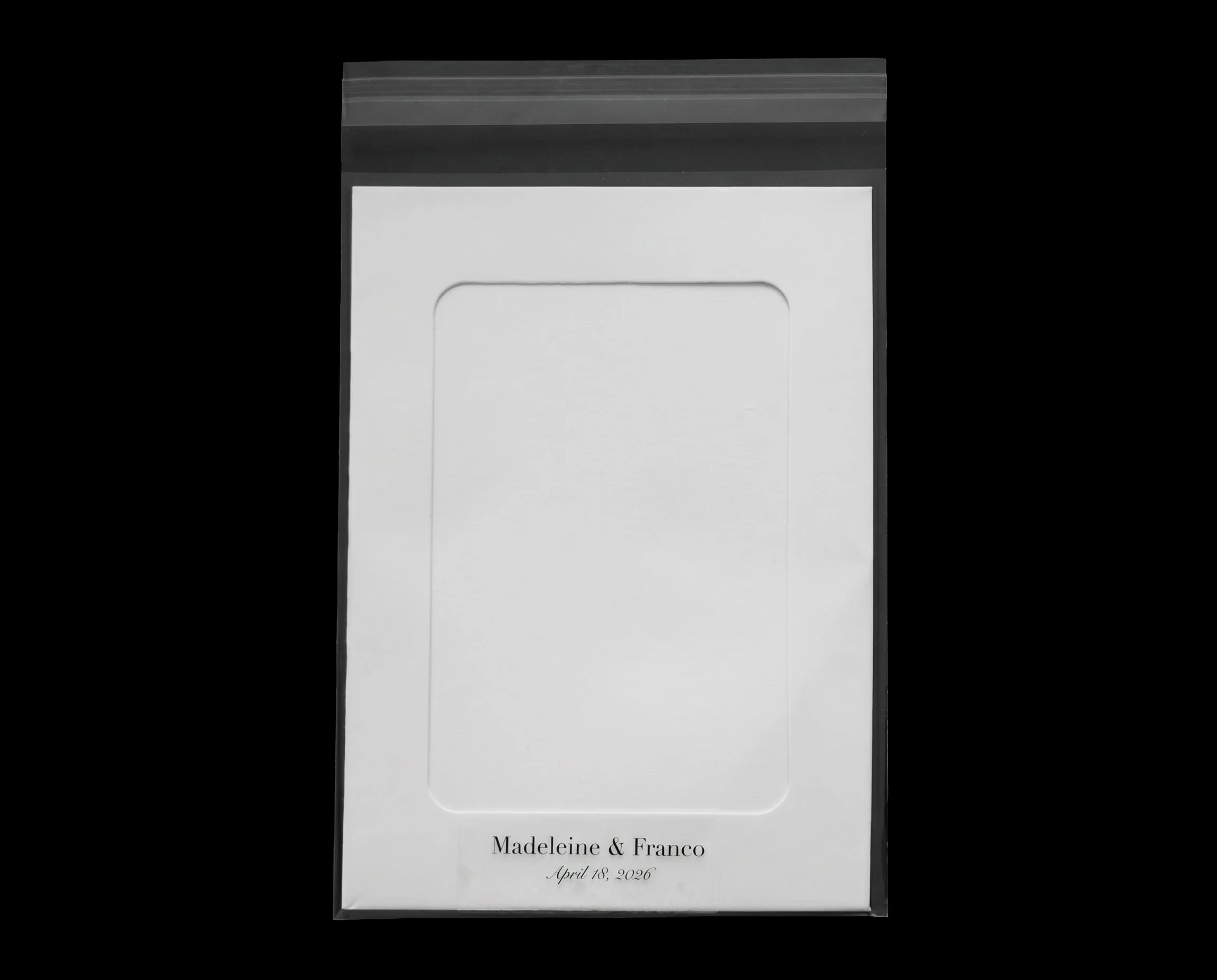 A customized white wedding invitation with black text that reads "Madeleine & Franco, April 18, 2026" and is enclosed in a clear plastic sleeve with a black top edge.