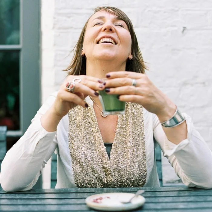 A woman smiling and enjoying a green beverage while sitting at an outdoor table.