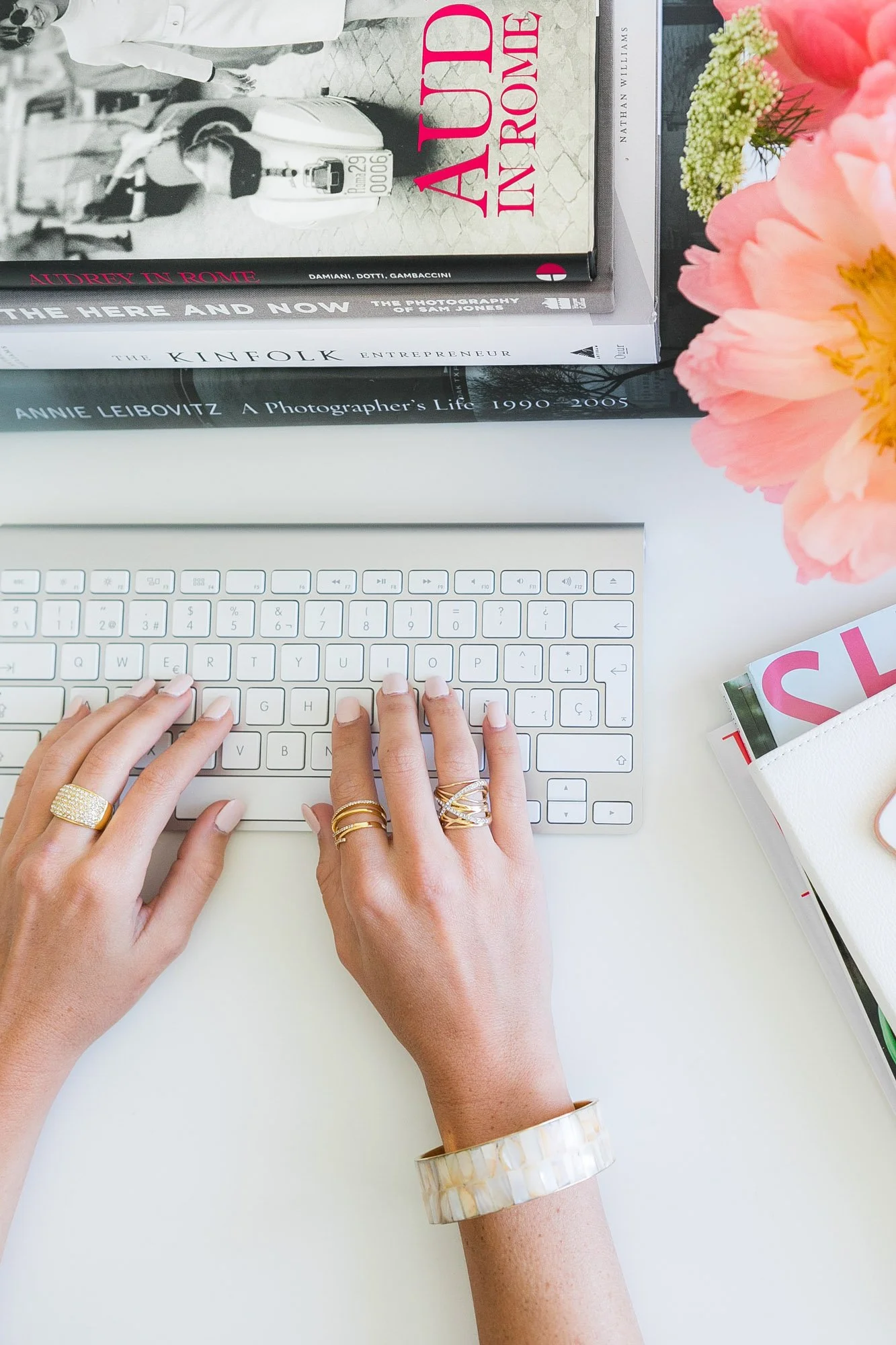 A person with multiple rings on their fingers and a large marble bracelet on their wrist is typing on a white keyboard. In the background, there are books stacked horizontally, with titles related to photography and individuals, and some flowers on the right side of the image.