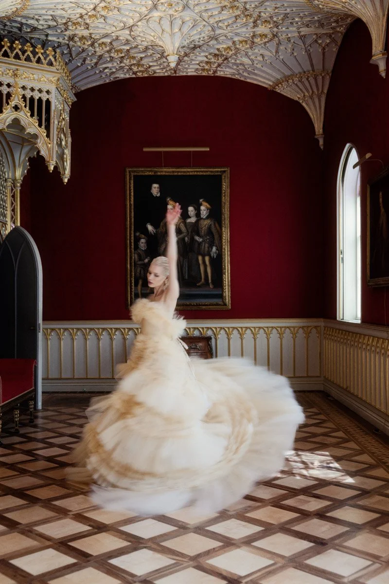 A woman in a white, feathered ball gown dances in an elegant, ornate room with gold detailing and red walls, illuminated by natural light from a window.