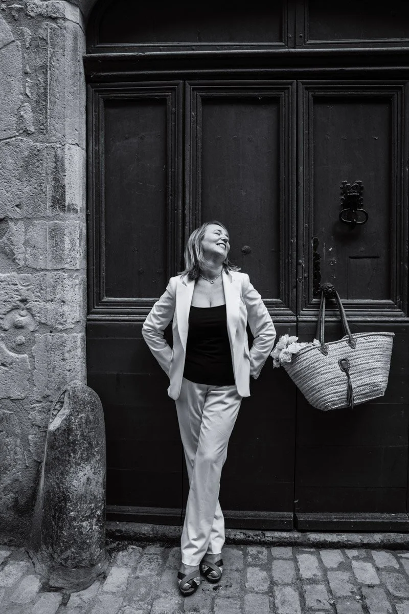 A woman in a light-colored suit and black top standing with hands behind her back, smiling and looking upward, in front of a large dark wooden door with a stone wall on the left and a bag hanging on the doorhandle.