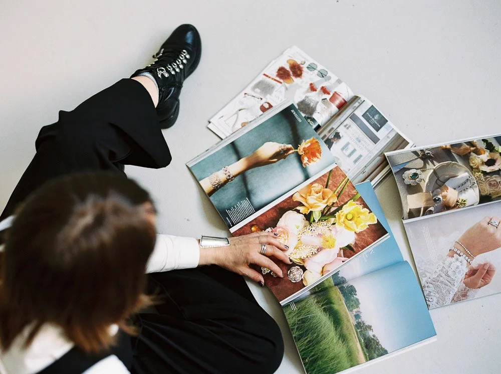 A person with brown hair and black shoes sitting on the floor looking at open magazines with images of flowers, jewelry, and outdoor scenery.