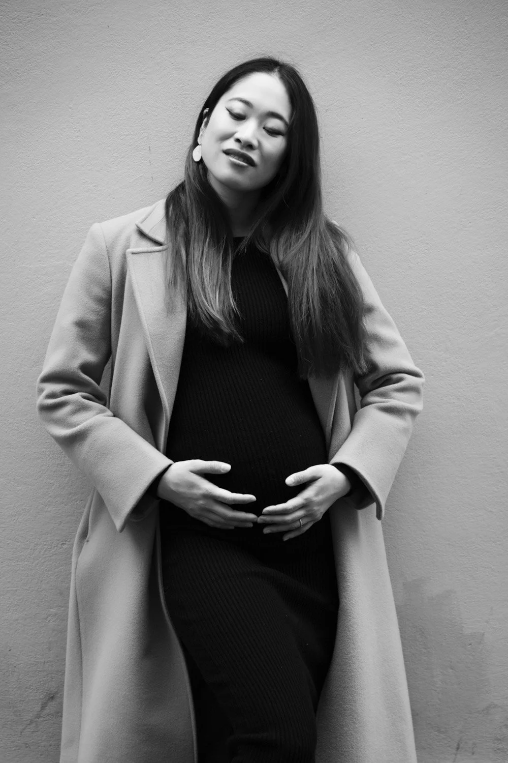 Black and white photo of a pregnant woman standing against a plain wall, with eyes closed and hands gently resting on her belly.