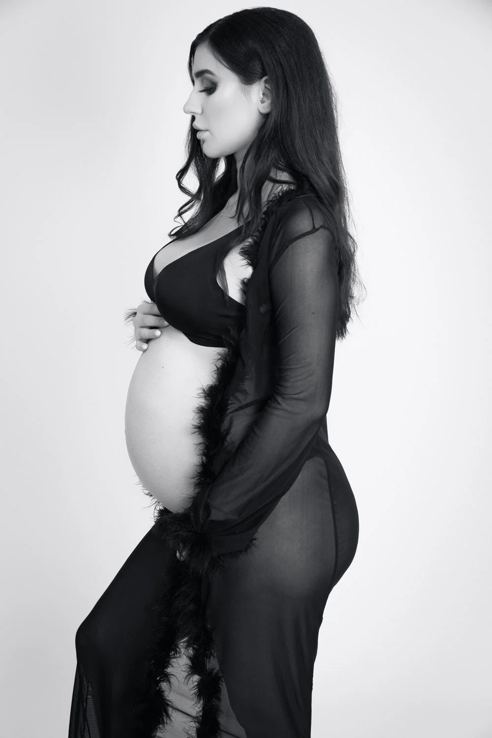 A pregnant woman with long dark hair, wearing a black bra and sheer black gown, standing in profile against a plain background.